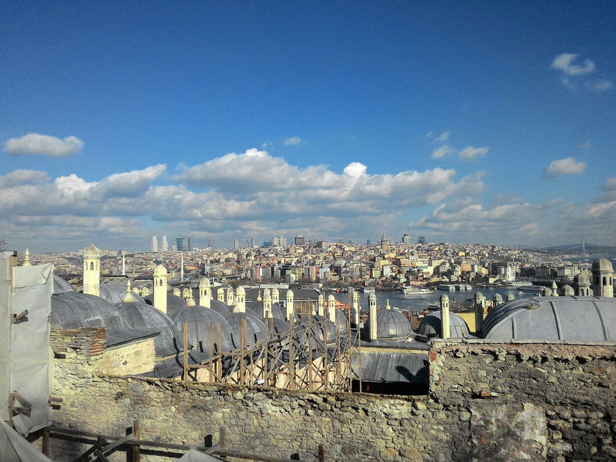 Панорама Стамбула с высоты: на переднем плане — плоские свинцовые купола мечети с дымниками, открытые в реставрацию; вдали — Золотой Рог, Галатская башня, деловой квартал Лейвент с небоскрёбами; яркое синее небо с облаками