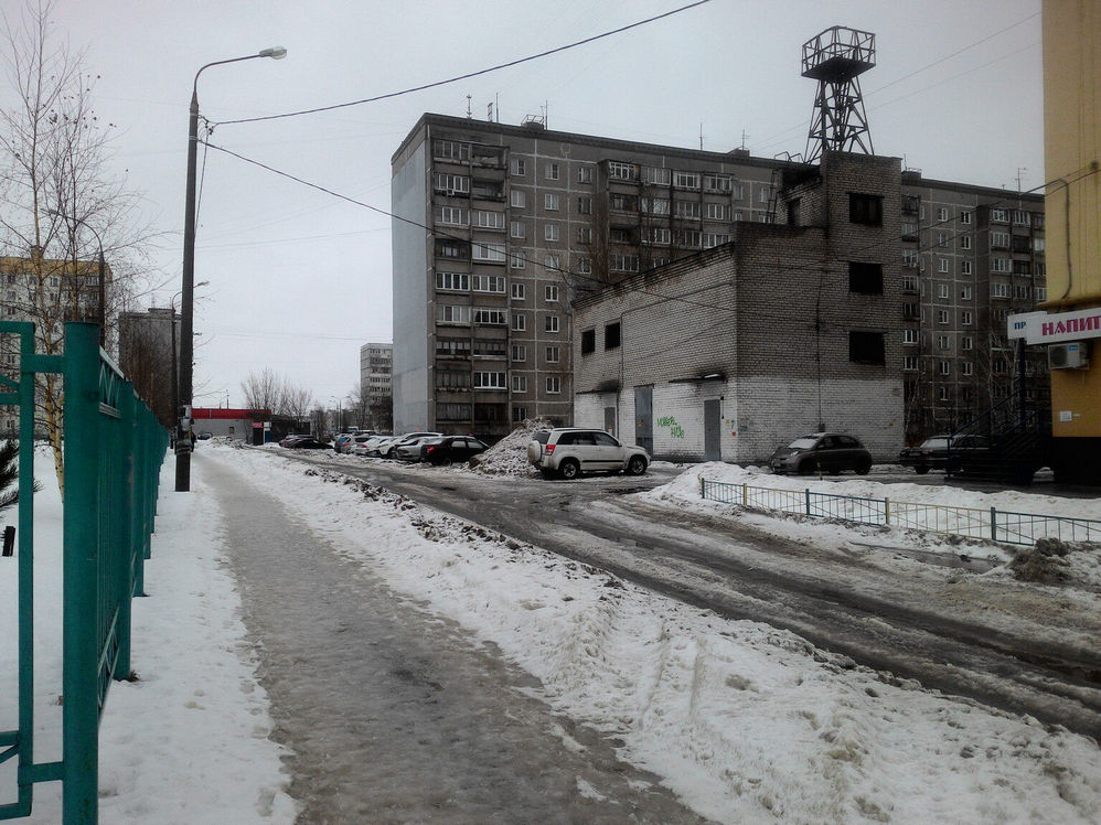 Фото зимней улицы: слева тротуар со снегом/слякотью и металлический зелёный забор; вдали 9-этажные панельные дома, небольшое одноэтажное строение; правее припаркованные машины и вывеска «НАПИ...» (Напитки?); над зданием видна высокая металлическая башня/вышка; пасмурное небо