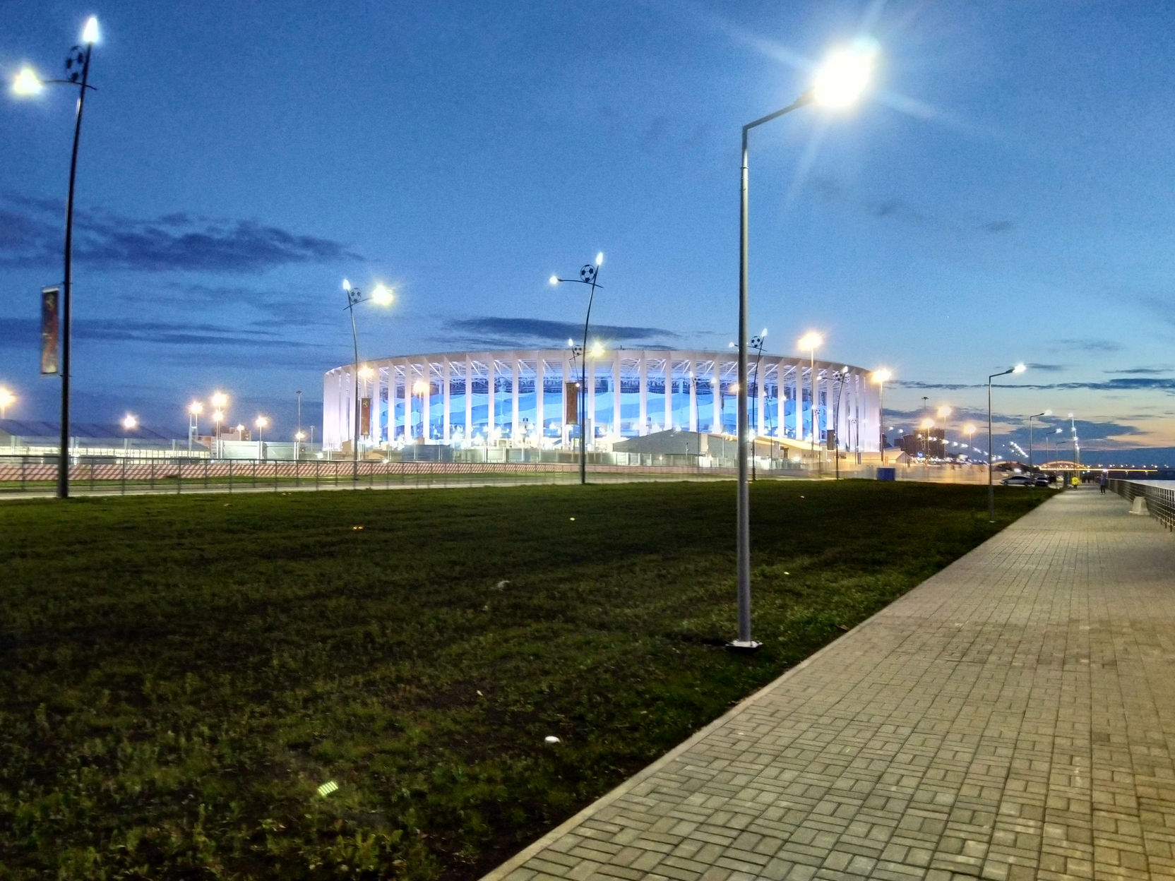 Ночная панорама: стадион «Нижний Новгород» (ЧМ-2018) в синей подсветке вдали; перед ним — газон, дорожки и фонари; голубое сумеречное небо