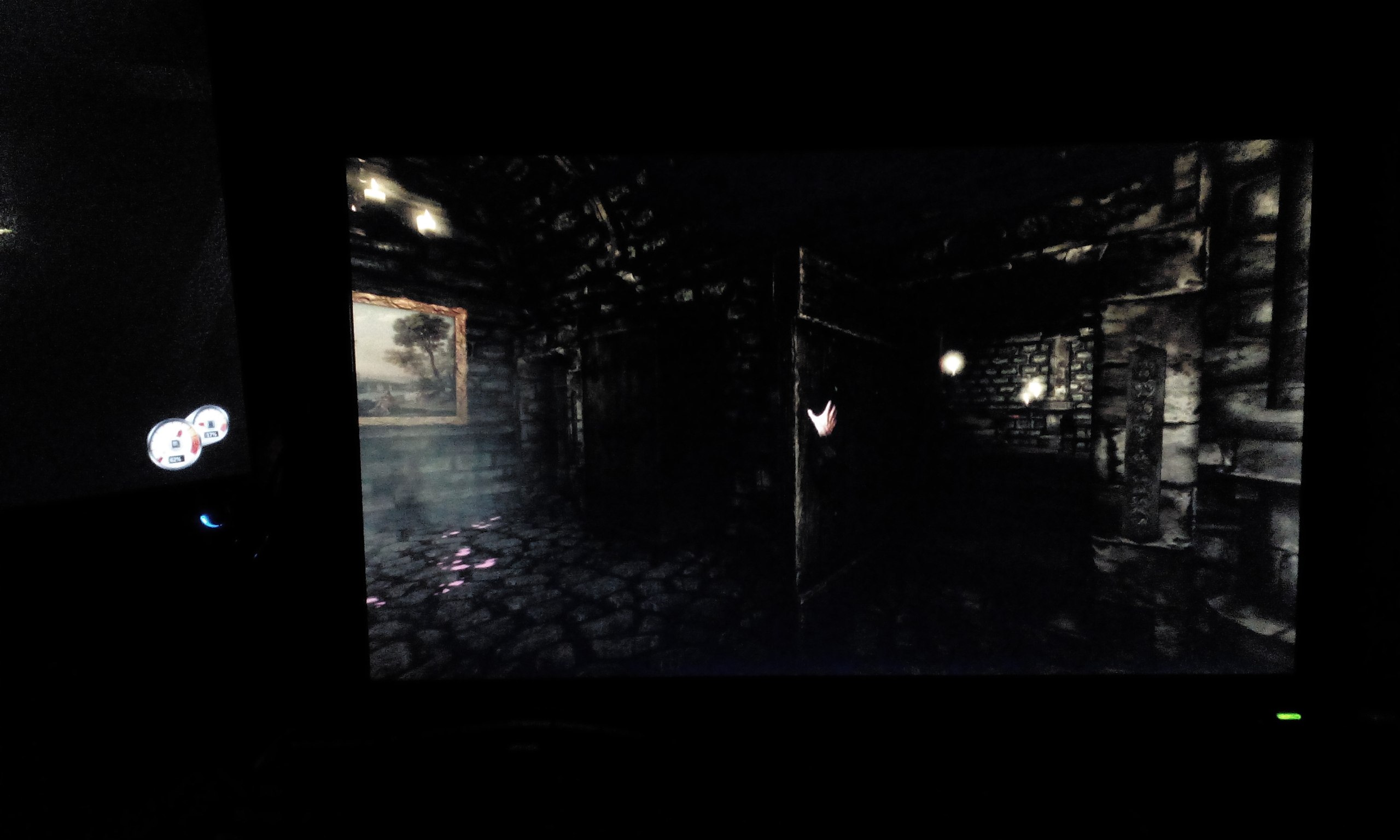 Монитор ПК в тёмной комнате: на экране игра Amnesia: The Dark Descent — тёмный коридор замка с каменными стенами, картина на стене, рука персонажа, одиночный фонарь; слева корпус ПК