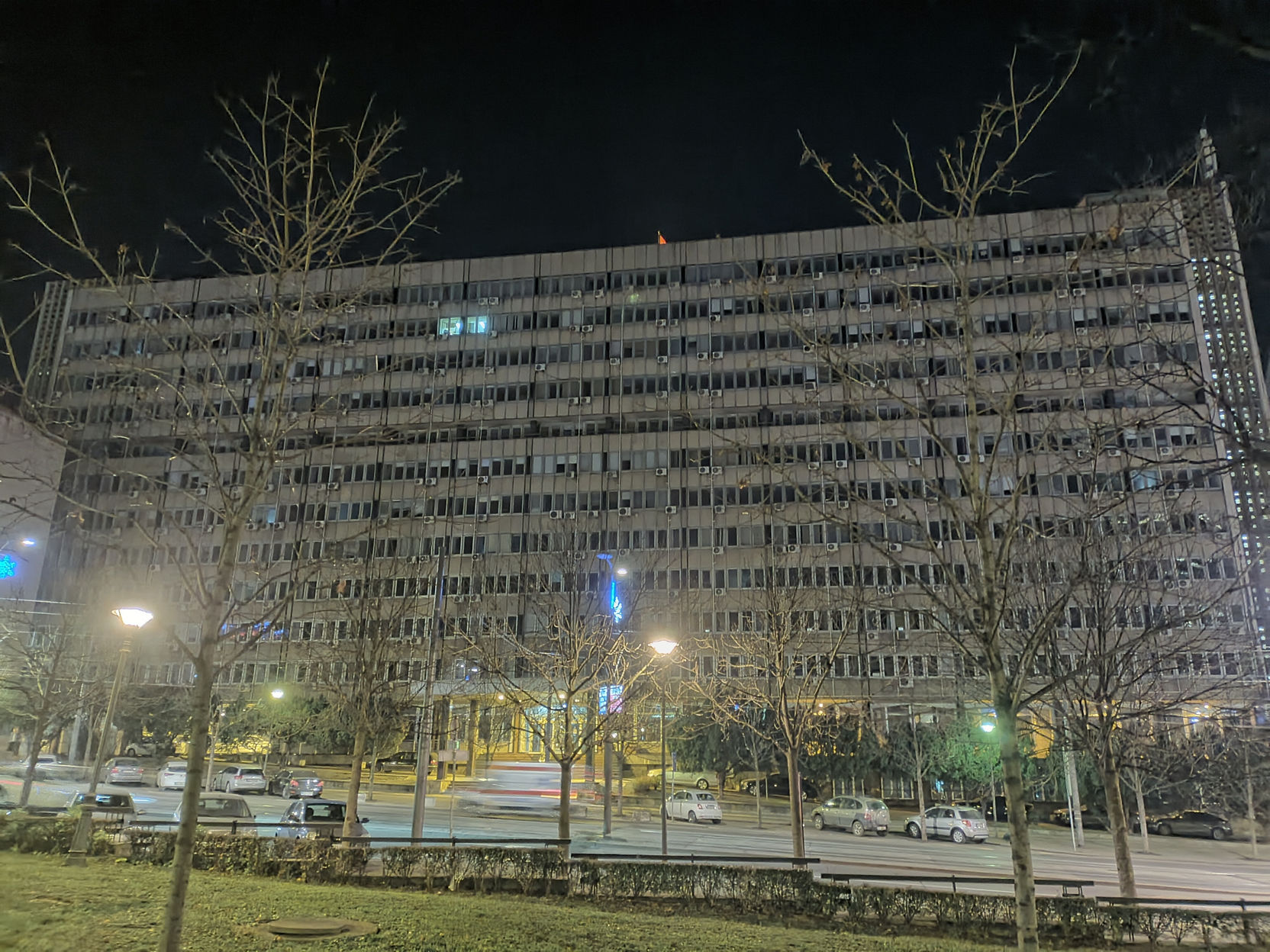 📷 Ночная фотография: большое многоэтажное административное здание прямоугольной формы с рядами окон; ночное освещение — фонари и светящиеся окна здания; на переднем плане широкая дорога с проезжающими автомобилями, голые деревья, газон.