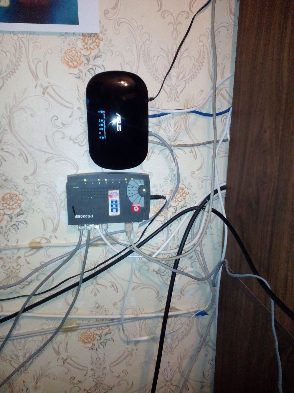 Роутер ASUS (чёрный) и свитч PS2208B, закреплённые на стене с обоями в цветочек; пучок кабелей по периметру