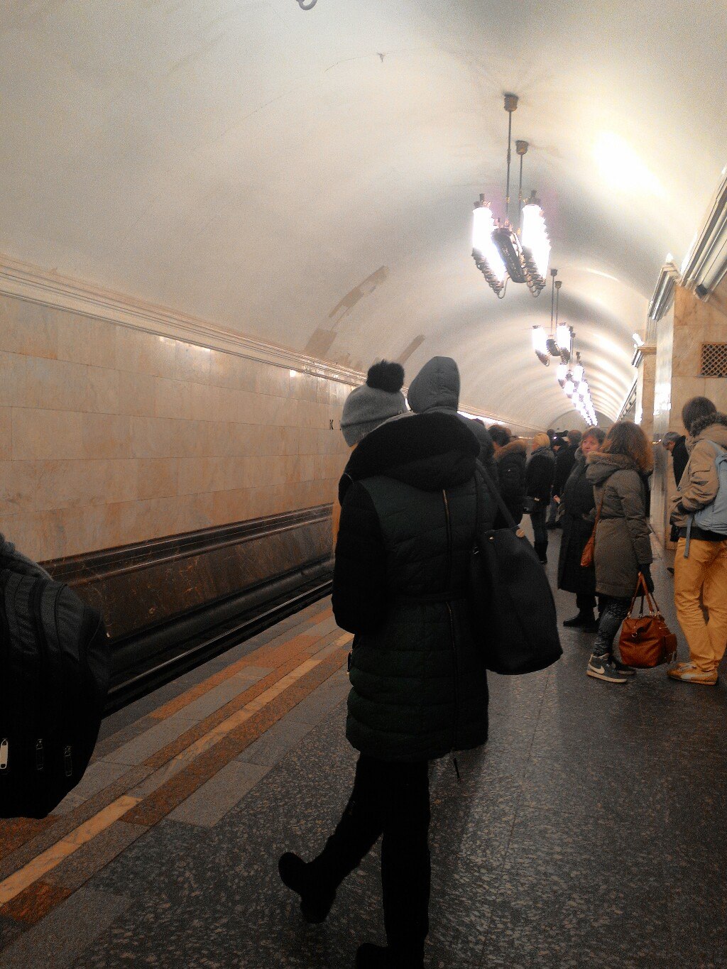 Платформа станции московского метро: высокий арочный свод из светлого мрамора, подвесные фигурные светильники-люстры; несколько пассажиров в зимних куртках стоят у края платформы; вдали — тоннель и ещё одна люстра
