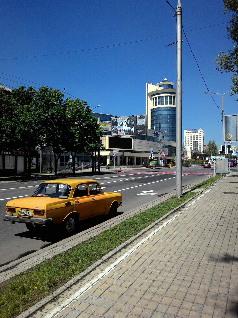 Улица Донецка в солнечный день: по дороге едет жёлтый Москвич-412; на заднем плане стеклянное здание с круглой башней (торговый центр); зелёные деревья, синее небо