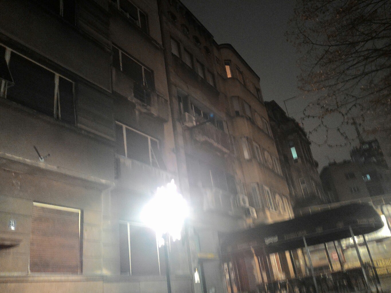 Ночной вид снизу на фасад белградского жилого дома: несколько этажей с балконами и тёмными окнами; внизу — ослепительно яркий декоративный фонарь-звезда; справа — силуэт автобуса или трамвая