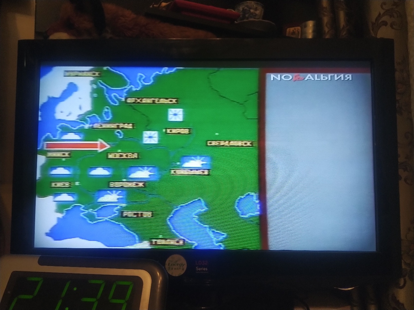 Телевизор, на экране канал «Ностальгия»: прогноз погоды с картой СССР — советские названия городов: Ленинград, Архангельск, Москва, Киров, Свердловск, Минск, Киев, Куйбышев, Воронеж, Ростов, Тюмень; красная стрелка указывает на Минск; внизу электронные часы 21:39