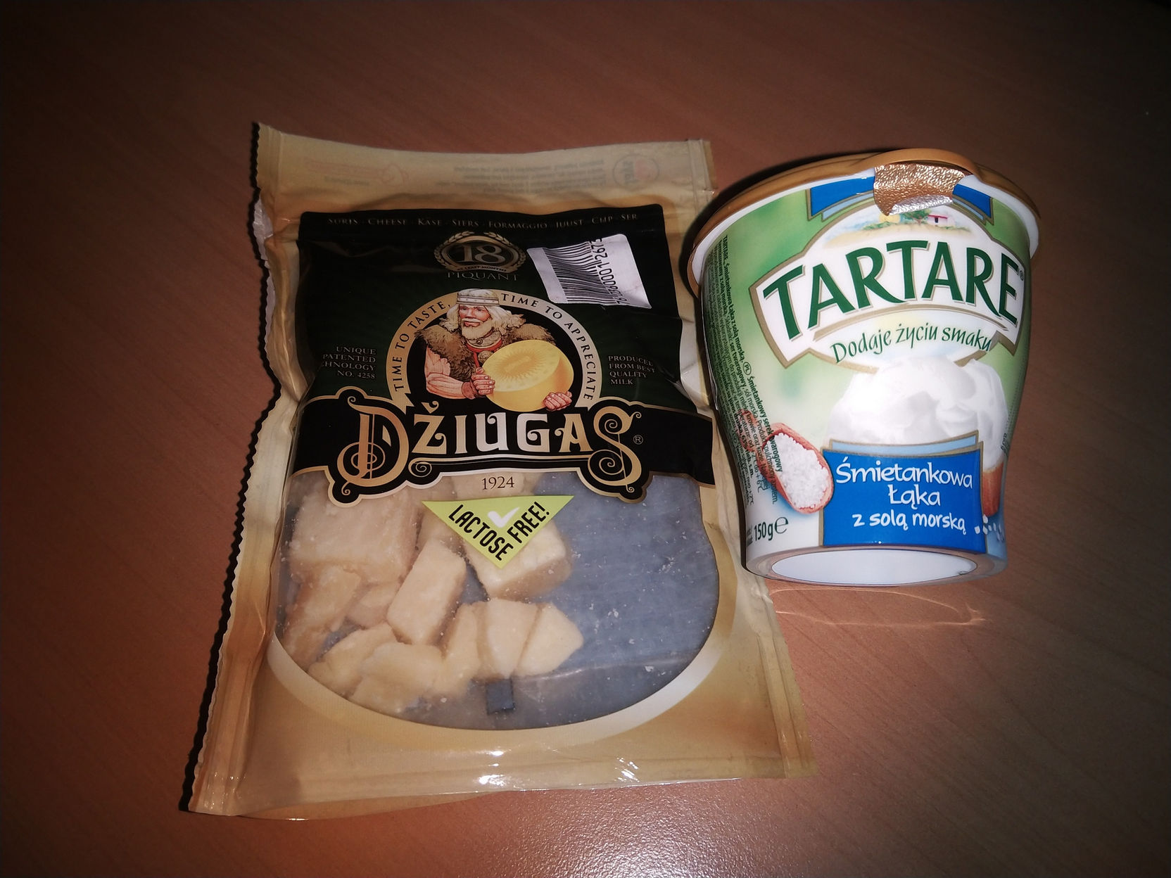 Два продукта на тёмной деревянной поверхности: слева — пакет литовского сыра «DŽIUGAS» с пометкой «LACTOSE FREE», внутри кусочки белого сыра; справа — пластиковый стакан польского соуса «TARTARE» с надписями «Dodaje życiu smaku» и «Śmietankowa Łąka z solą morską»