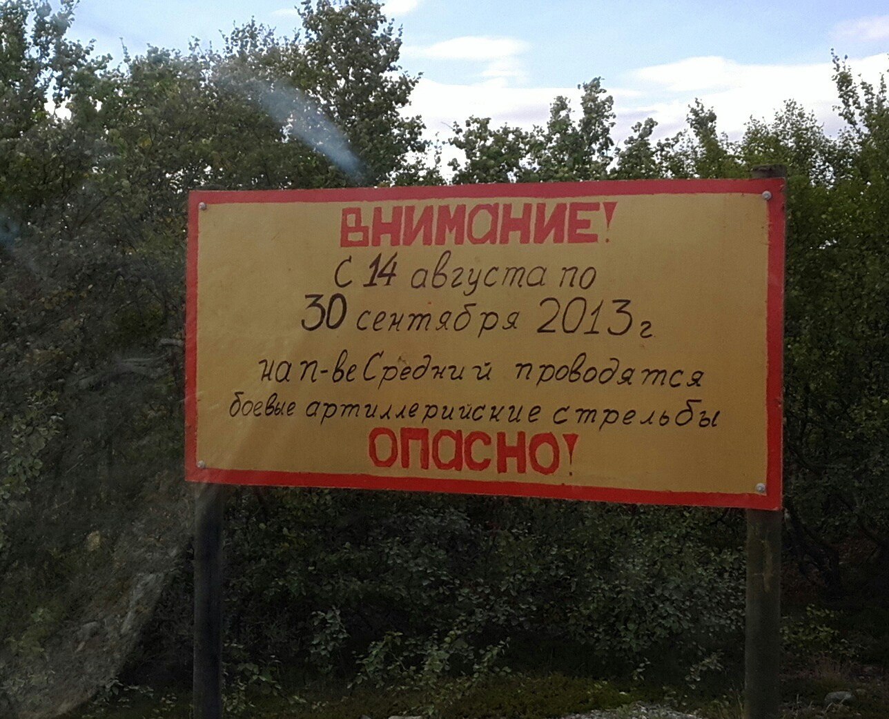 Жёлтый прямоугольный предупреждающий знак у лесной дороги: красным напечатано «ВНИМАНИЕ!» и «ОПАСНО!», чёрным — «С 14 августа по 30 сентября 2013 г. на п-вСредний проводятся боевые артиллерийские стрельбы»; на фоне деревьев и голубого неба