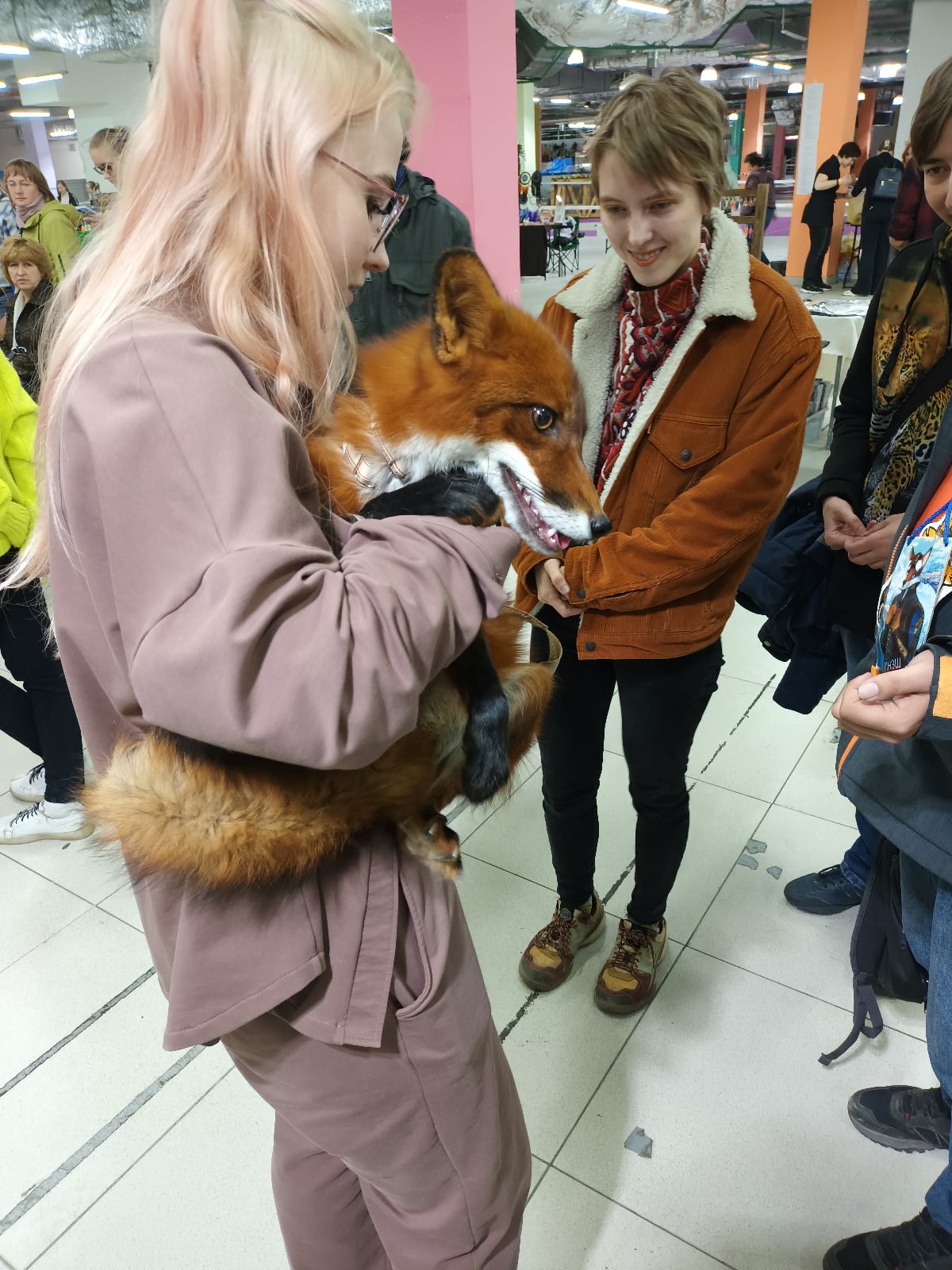 📷 Женщина со светлыми/платиновыми волосами в бежевом наряде держит рыжую лису; рядом человек в коричневой куртке; торговый центр.