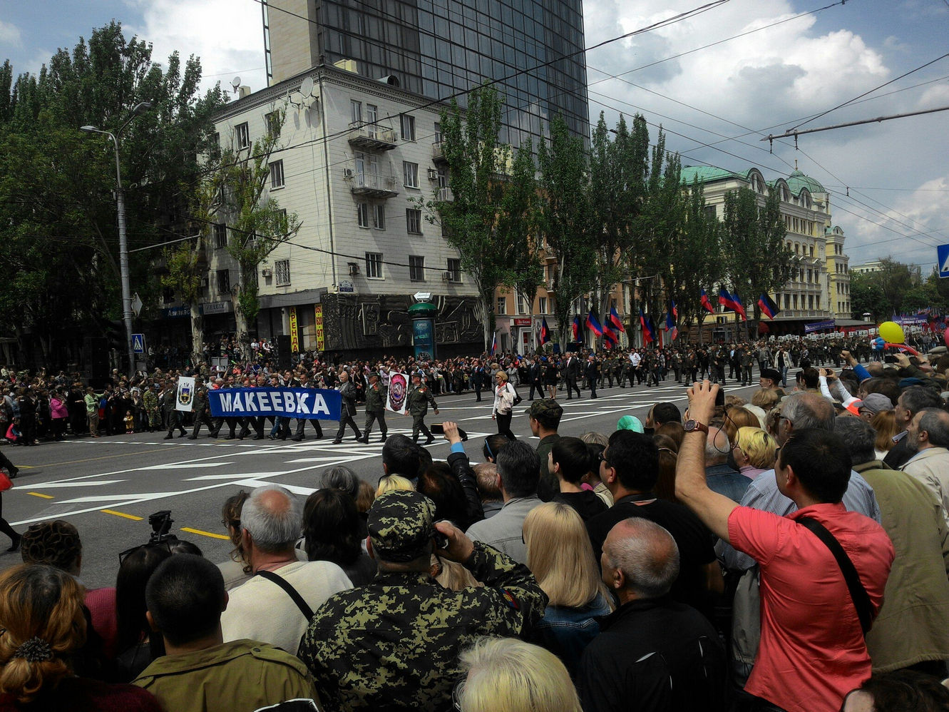 Парад колонны с транспарантом «МАКЕЕВКА» по центральному проспекту Донецка: участники в тёмной одежде, флаги ДНР; зрители на тротуаре снимают на телефоны; на заднем плане стеклянная высотка