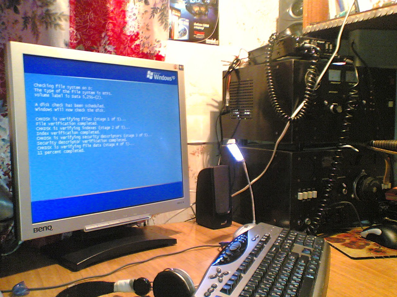 Фото рабочего стола: монитор BenQ с синим экраном (установка Windows или BSOD с текстом процесса проверки файлов); справа радиостанция/трансивер; USB-фонарик; клавиатура; на стенах постеры