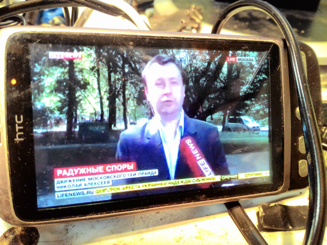 HTC смартфон: LifeNews, LIVE Москва; заголовок «РАДУЖНЫЕ СПОРЫ»; подпись «Движение московского гей-прайда Николай Алексеев»; бегущая строка «lifenews.ru ...СРОК АРЕСТА УКРАИНКИ НАДЕЖДЫ СА... СРОЧНО СПУТНИК...»