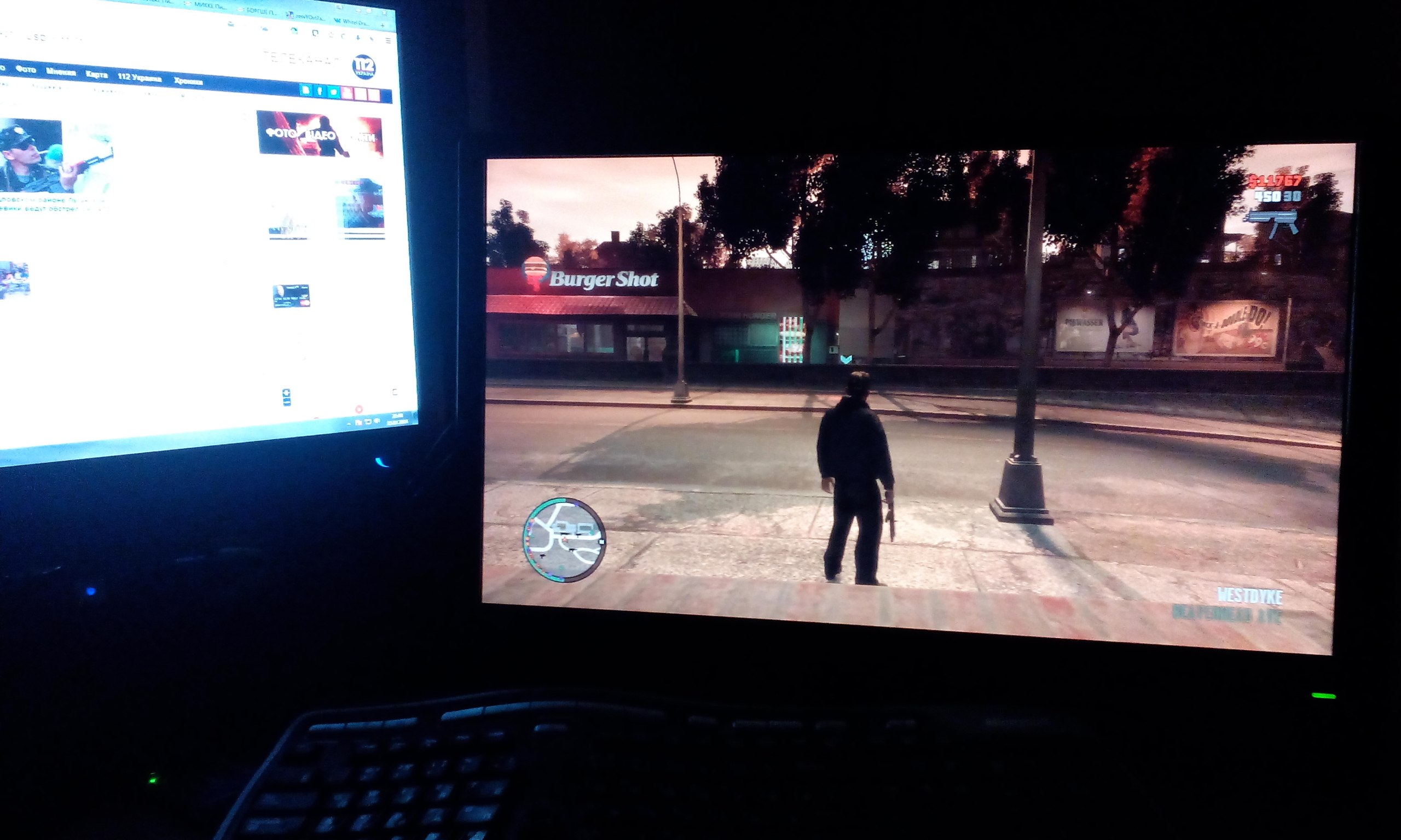 Два монитора в тёмной комнате: левый — новостной сайт «112 Украина»; правый — GTA IV: персонаж стоит у Burger Shot в районе Westdyke