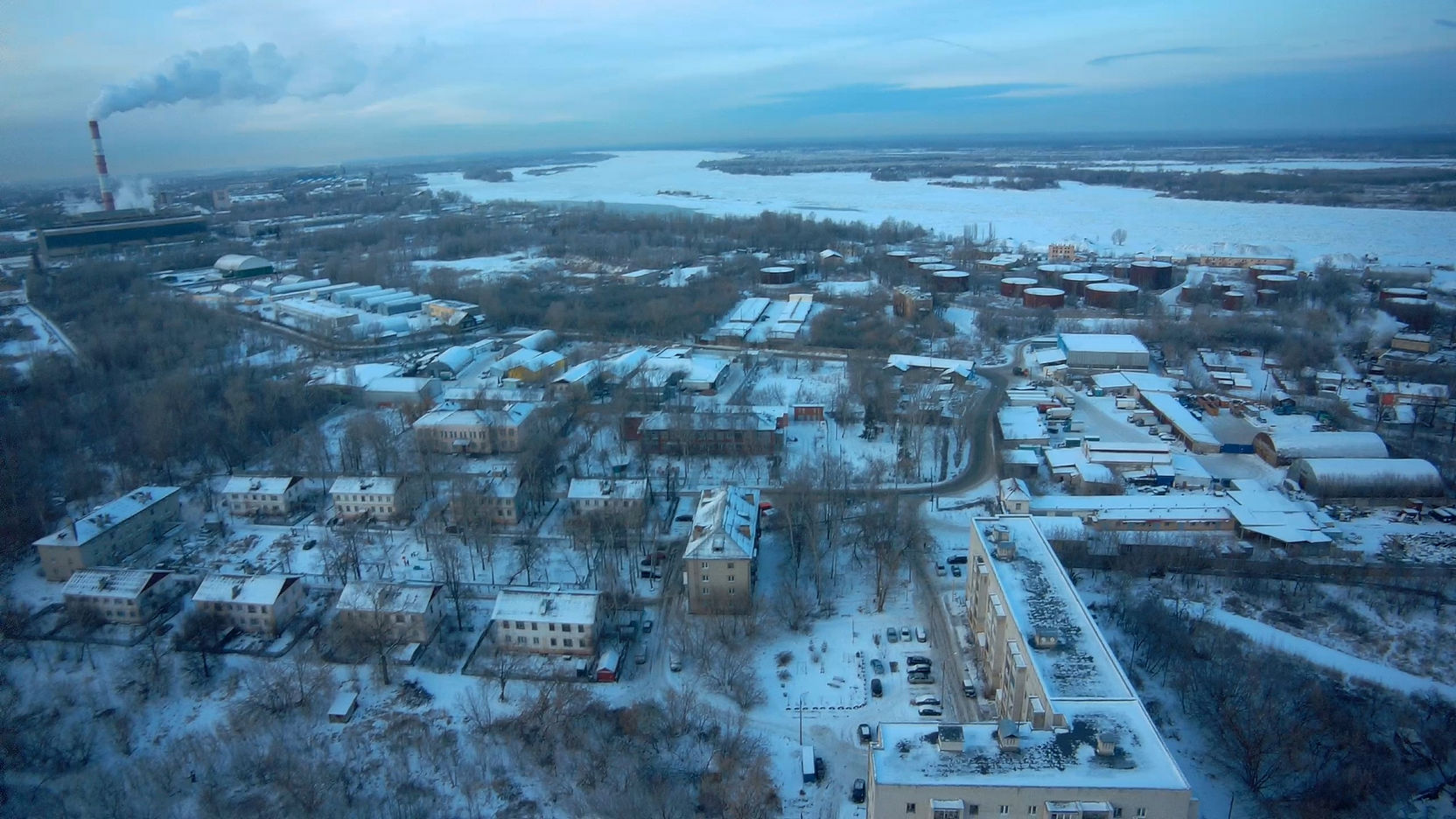 📷 Аэросъёмка заснеженного города: жилые кварталы и промзона, замёрзшая река/водохранилище, дымящая труба котельной, голубое небо с облаками