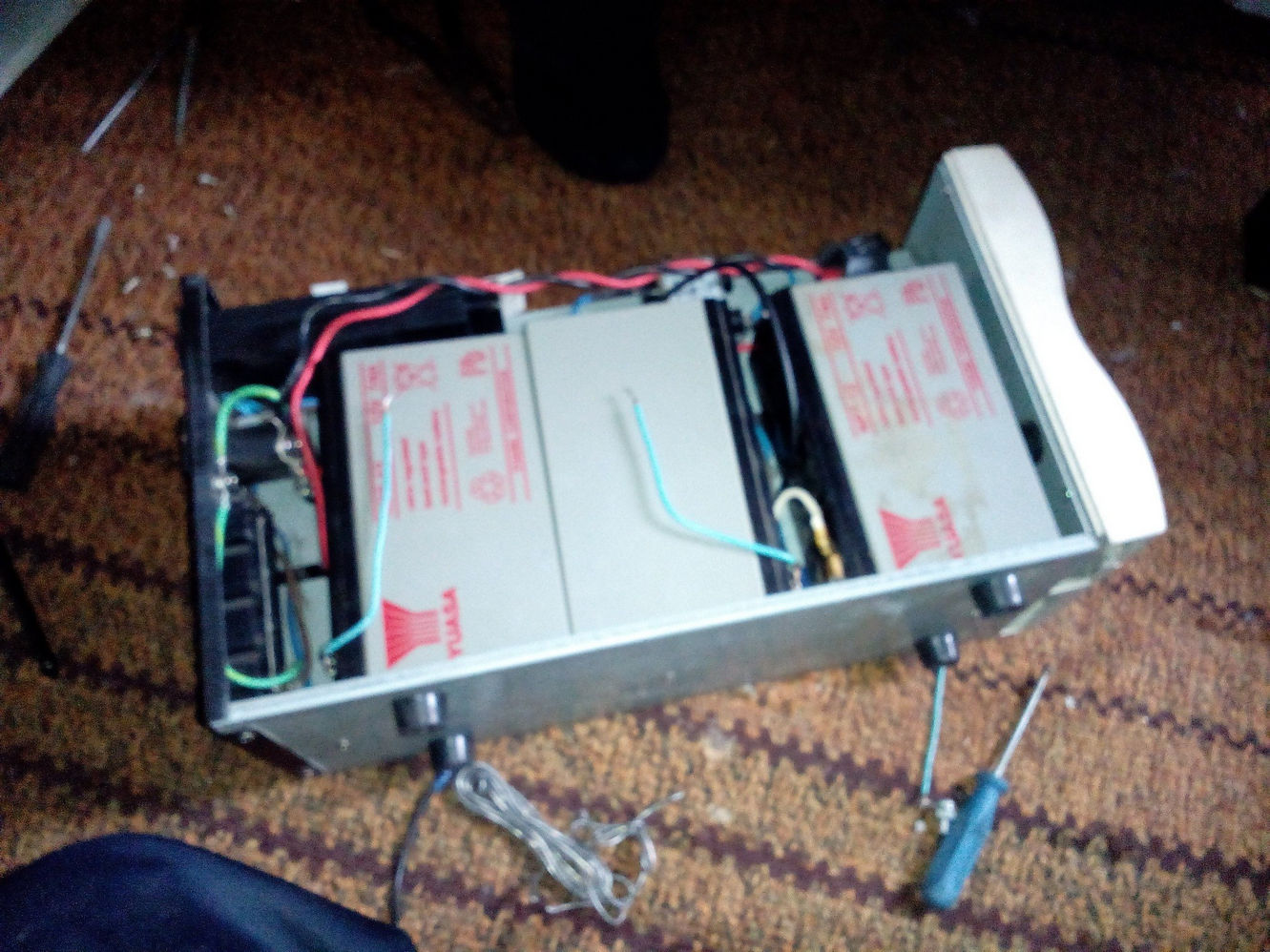 Разобранный ИБП APC Smart-UPS 600: открытый нижний отсек с двумя аккумуляторами YUASA, красными и синими проводами; рядом отвёртка