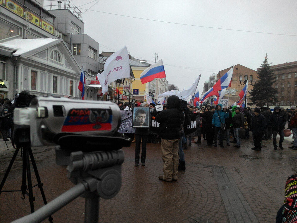Фото уличного митинга: на переднем плане камера на штативе с наклейками, вдали колонна людей с российскими триколорами и плакатами, на одном из них портрет Немцова; пасмурная зимняя погода, городская улица