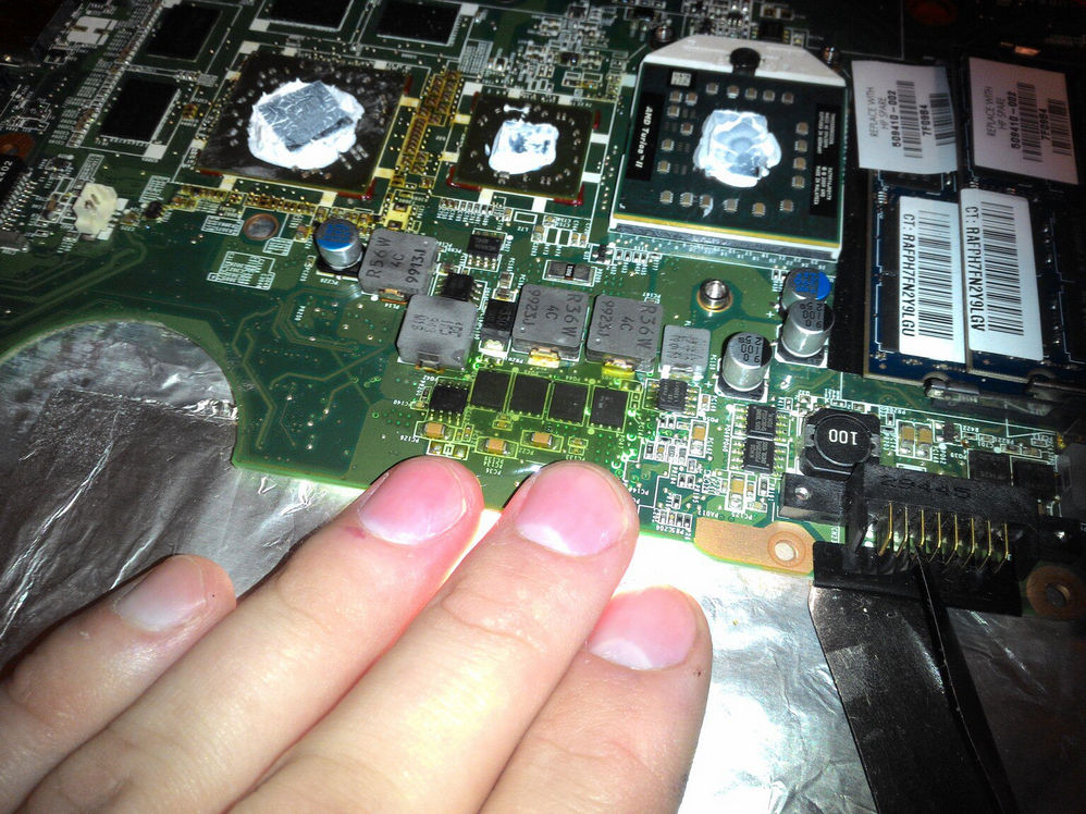 Крупный план материнской платы ноутбука: три BGA-чипа (GPU и мосты) покрыты термопастой и обёрнуты фольгой для рефлоу; рука придерживает плату снизу; справа стикер с серийным номером