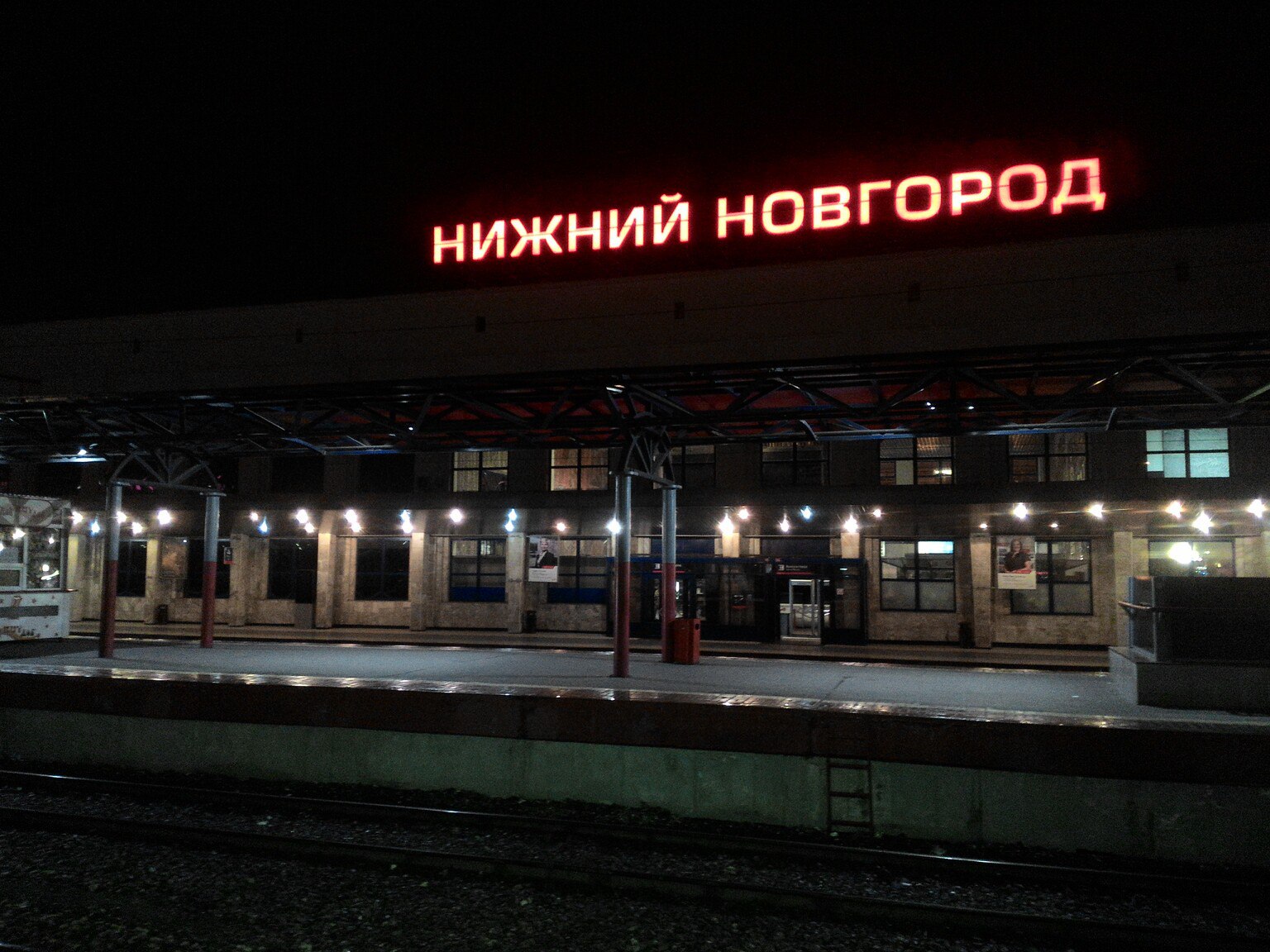 Ночь: здание железнодорожного вокзала с красной неоновой надписью «НИЖНИЙ НОВГОРОД» над крышей; вдоль перрона красные металлические опоры с гирляндами ламп; два этажа здания, тёмное небо