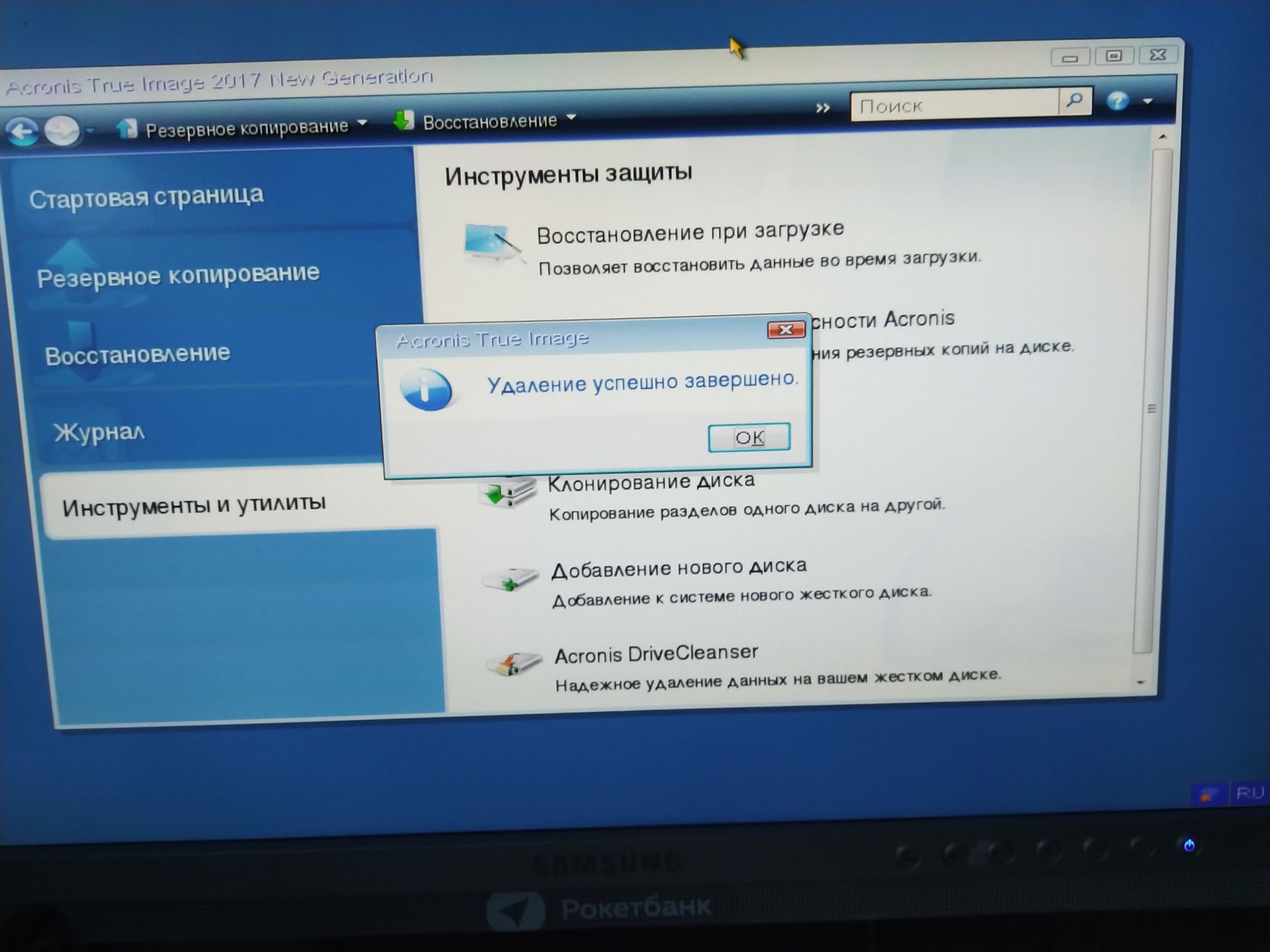 Скриншот Acronis True Image 2013 (Next Generation): левое меню — «Стартовая страница», «Резервное копирование», «Восстановление», «Журнал», «Инструменты и утилиты»; страница «Инструменты защиты» — «Восстановление при загрузке», «Клонирование диска», «Добавление нового диска», «Acronis DriveCleanser — Надёжное удаление данных на жёстком диске»; поверх — всплывающий диалог «Удаление успешно завершено.» с кнопкой «ОК»; внизу логотип «РокетБанк»