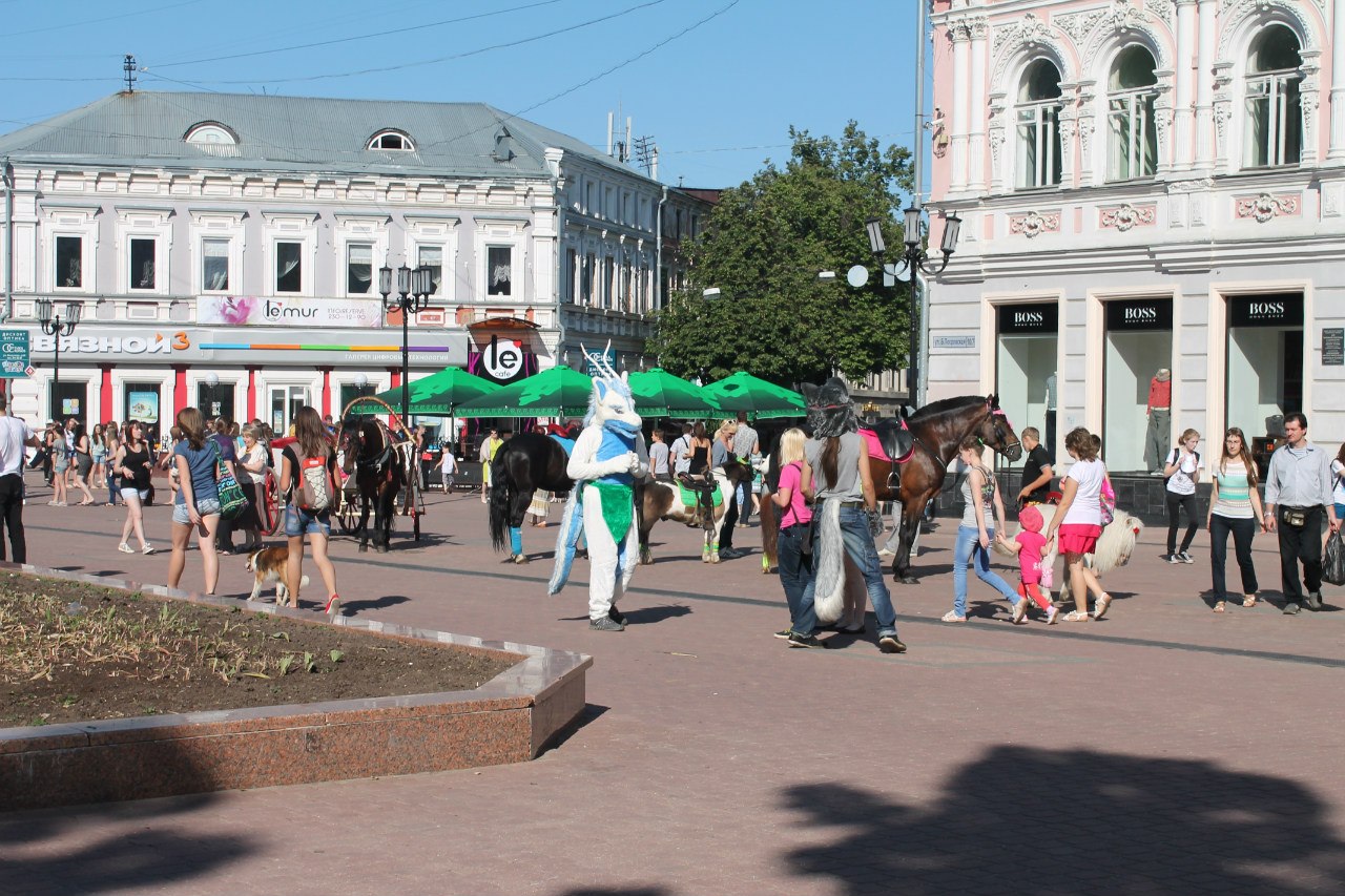 Пешеходная улица Нижнего Новгорода: человек в костюме синего дракона среди прохожих, рядом ведут лошадей, вдали магазины le'mur и Boss
