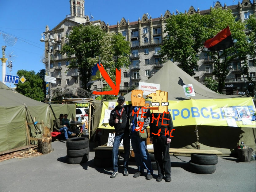 📷 Трое стоят у палаточного лагеря на Майдане Незалежности в Киеве: левый — в маске лисы и тёмной одежде, средний — с бумажной маской сбоку, правый — в обычной одежде; красная стрелка вниз указывает на левого; за ними палатки, баннер «...КРОВСЬКИХ...»; на заднем плане здания, флаги.