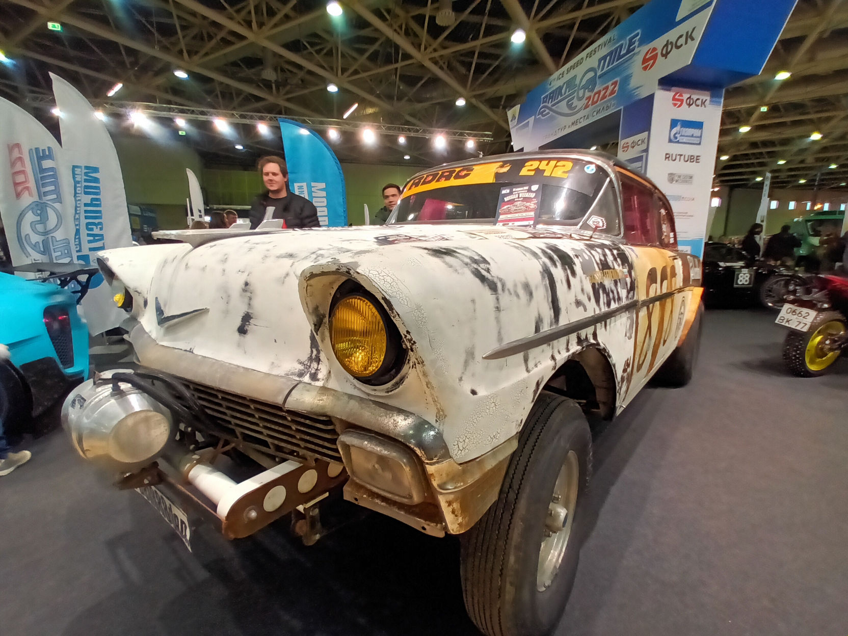 📷 Выставка: кастомный ретро-автомобиль 1950-х в стиле рэтрод — белый с ржавчиной, надпись «STRONG # 242»; двое людей рядом; баннеры «SUTURE», «ФСК», «ГАЗПРОМ».
