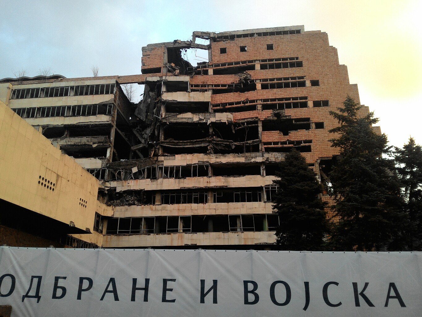 Разрушенное здание Генерального штаба МО Сербии в Белграде — последствия авиаударов НАТО 1999 года: выгоревшие перекрытия, обнажённые балки, выбитые окна; внизу забор с надписью «ОДБРАНЕ И ВОЈСКА»