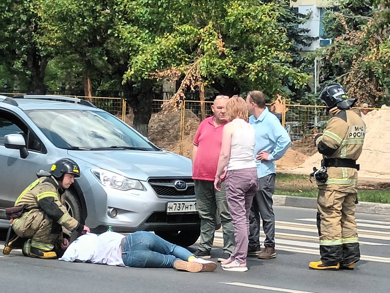📷 Место ДТП на пешеходном переходе: пострадавший в джинсах и розовых кроссовках лежит на асфальте; рядом — серебристый Subaru (номер Р737ВМ); двое пожарных в форме (жёлто-оранжевые куртки) и двое гражданских; деревья на заднем плане.