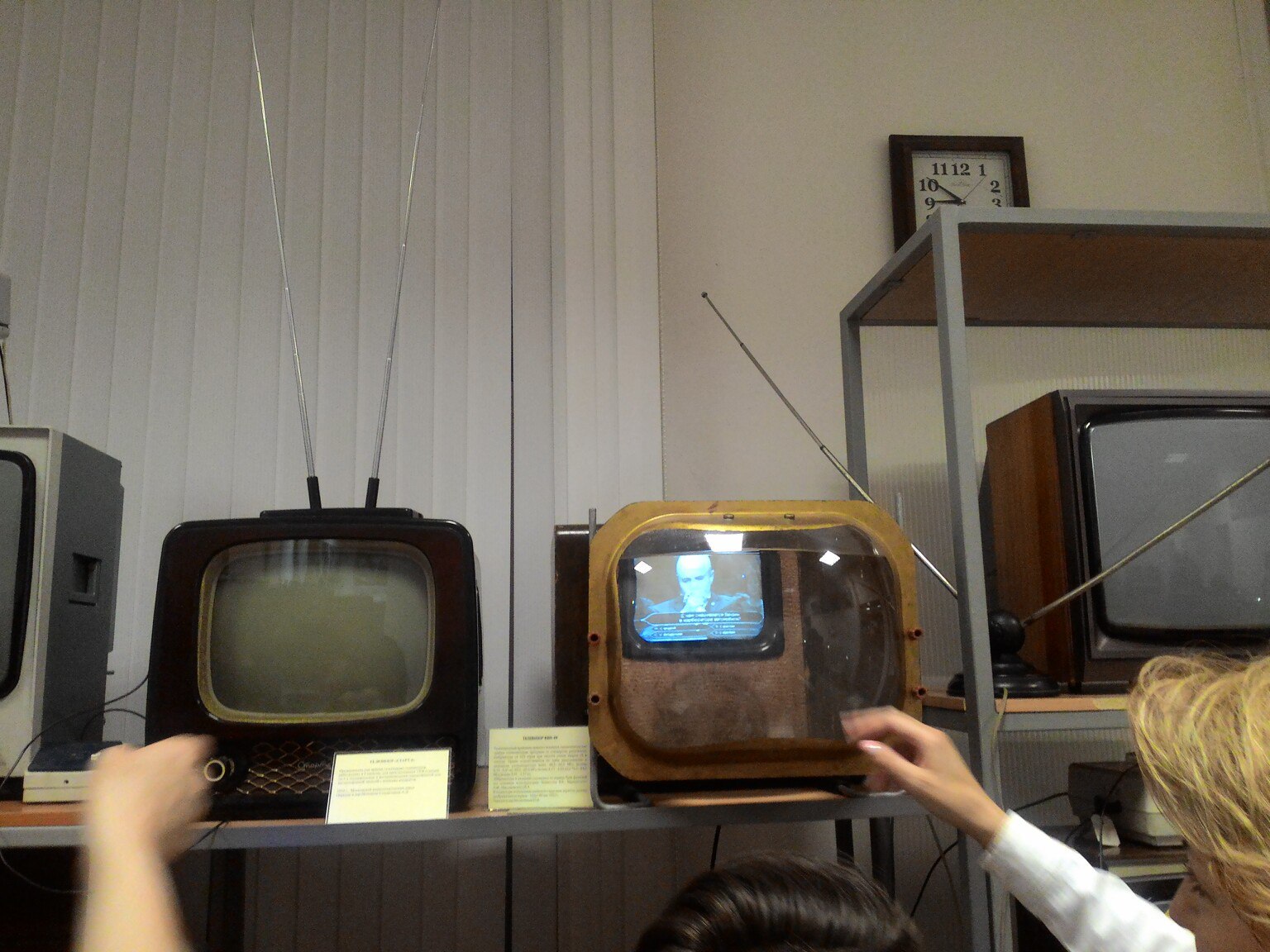Экспозиция музея: три ранних советских телевизора 1950–60-х гг. на полках — слева чёрный прямоугольный (с экраном без изображения), по центру деревянный в форме тумбы (на экране чёрно-белое изображение человека), справа ещё один; руки посетителей; на стене в правом верхнем углу — настенные часы