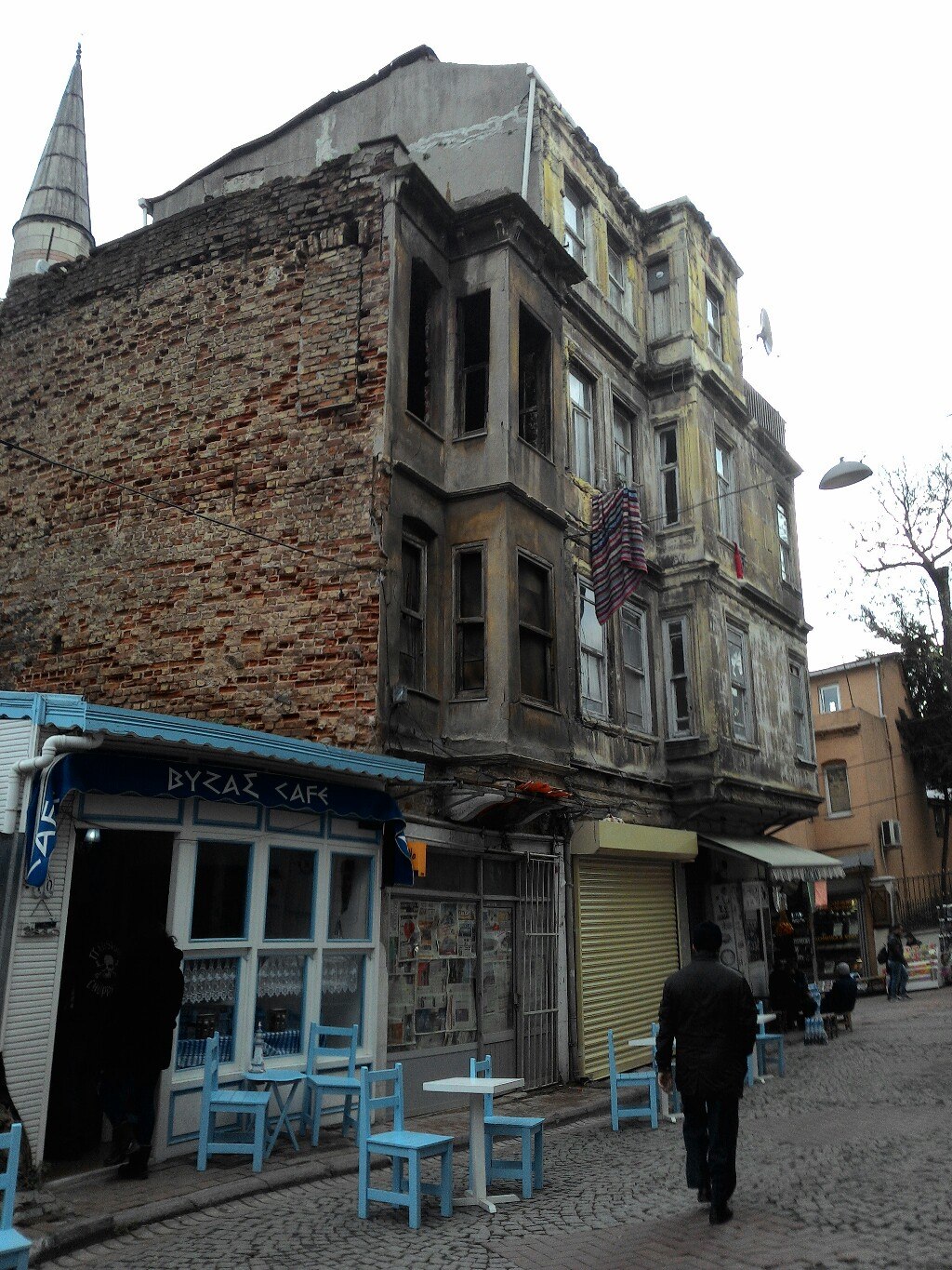 Угловой ветхий дом Стамбула: внизу — кафе «BYZAS CAFE» с синими стульями; рядом — торец средневековой кирпичной стены и минарет мечети; прохожий; мощёный тротуар
