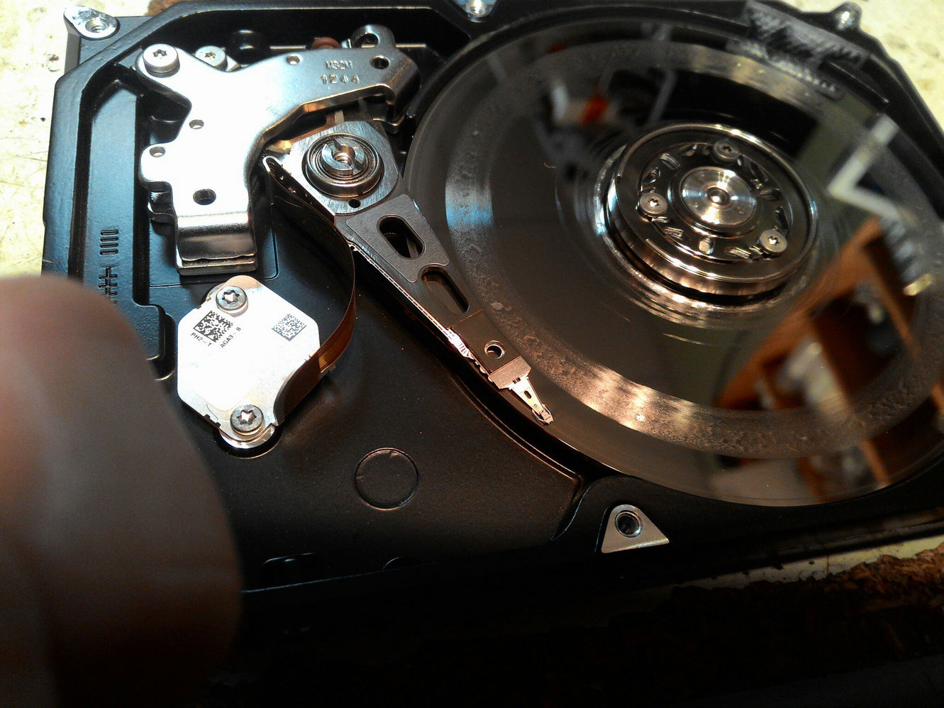 Крупный план вскрытого жёсткого диска (HDD): блестящие магнитные пластины справа, рычаг головки чтения/записи с тонким наконечником, наклейка с QR-кодом и надписью «RH1 04×3 B» на актуаторе, ось шпинделя с креплениями; корпус диска тёмный металлический