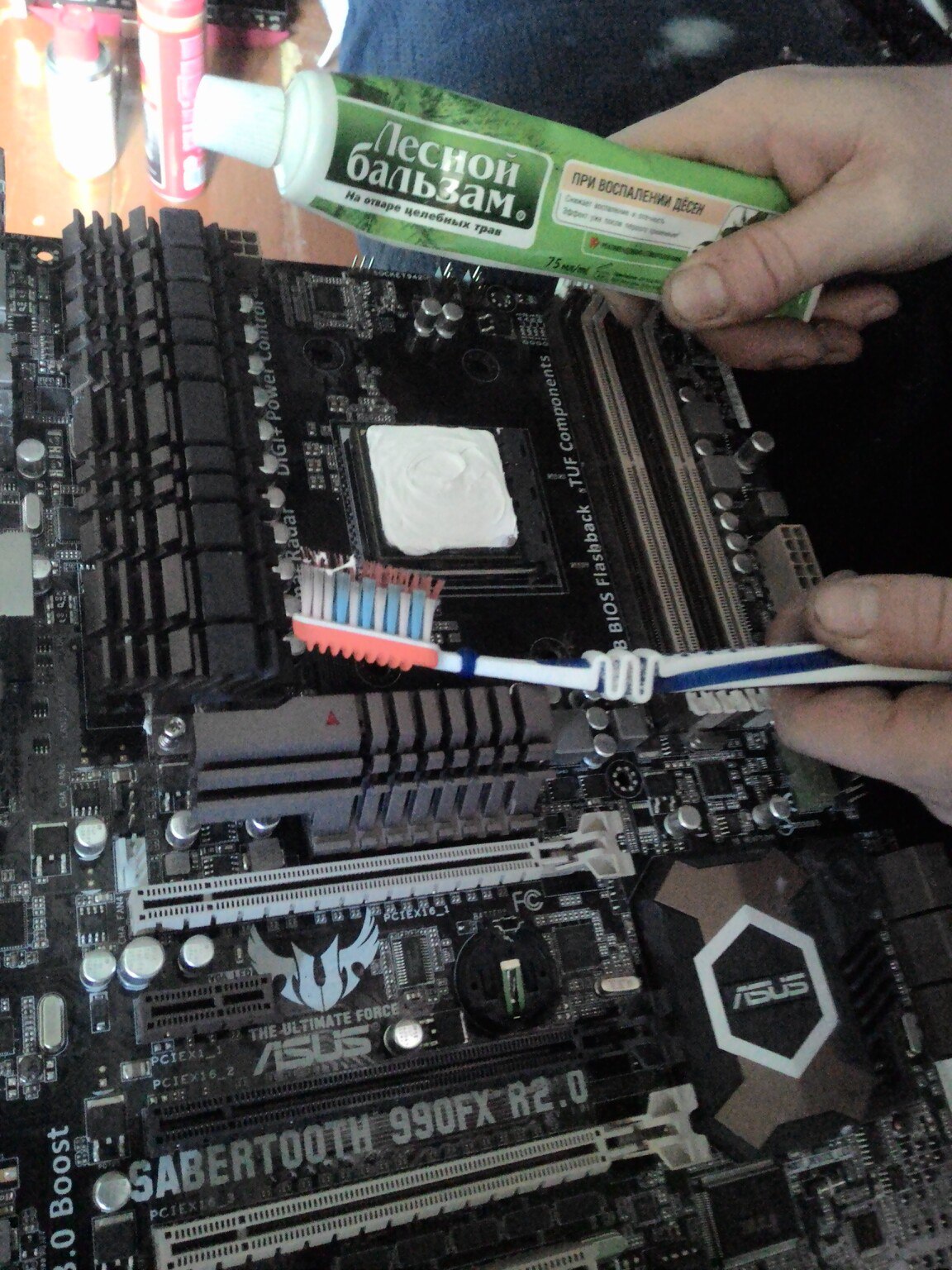 Нанесение термопасты на процессор материнской платы ASUS SABERTOOTH 990FX R2.0: рука держит тюбик «Лесной бальзам» и зубную щётку; на CPU-сокете белая термопаста; надписи на плате: DIGI+ Power Control, BIOS Flashback, TUF Components, ASUS