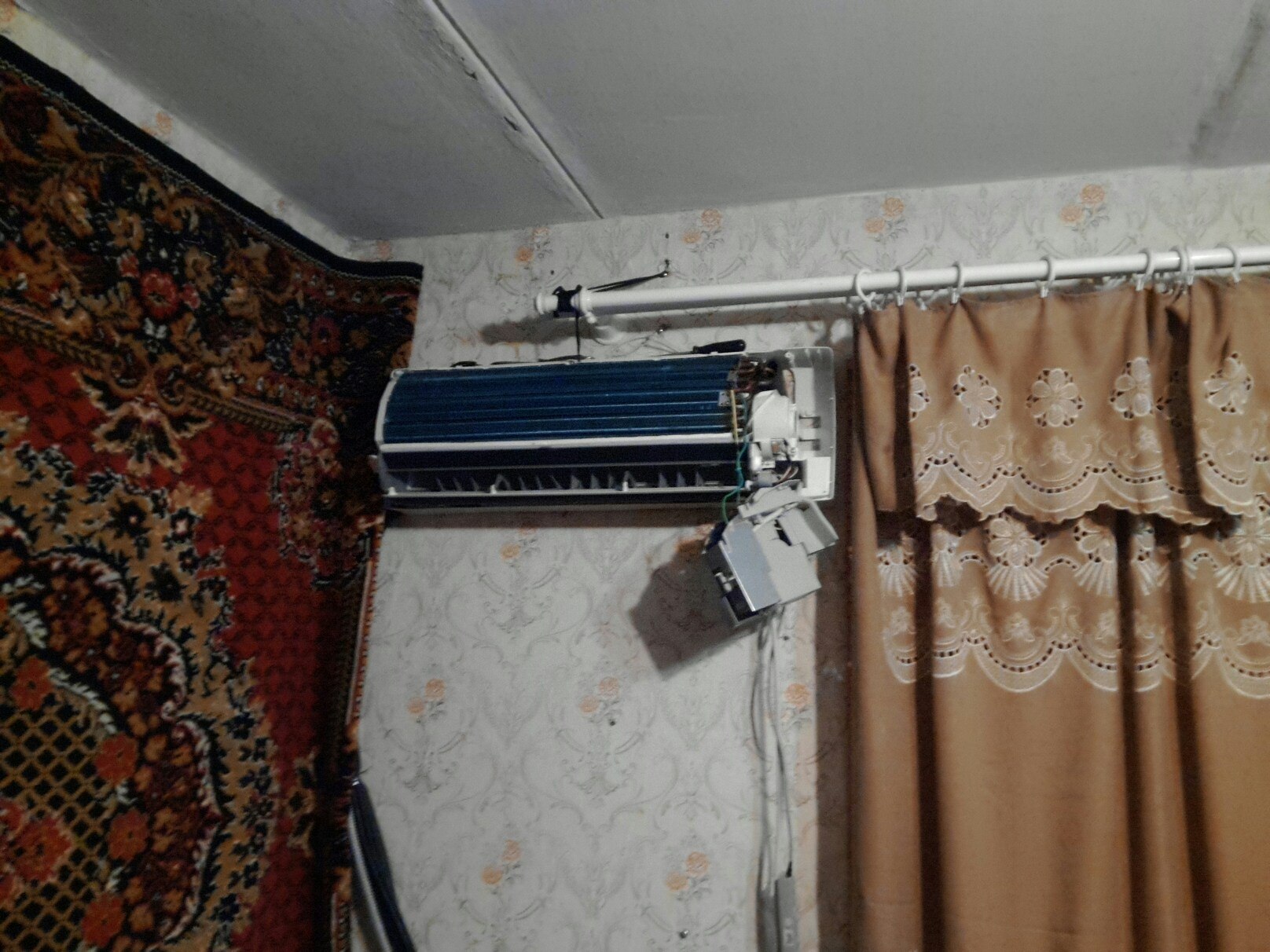 Разобранный внутренний блок кондиционера на стене в комнате с цветчатыми обоями, рядом ковёр и шторы