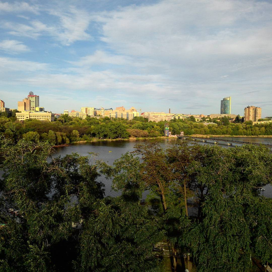 Панорама города с высоты: широкая река или водоём с отражением неба, густые кроны зелёных деревьев по берегам, вдали многоэтажные жилые дома (5–16 этажей) и высотные здания; голубое небо с лёгкими облаками, летний день