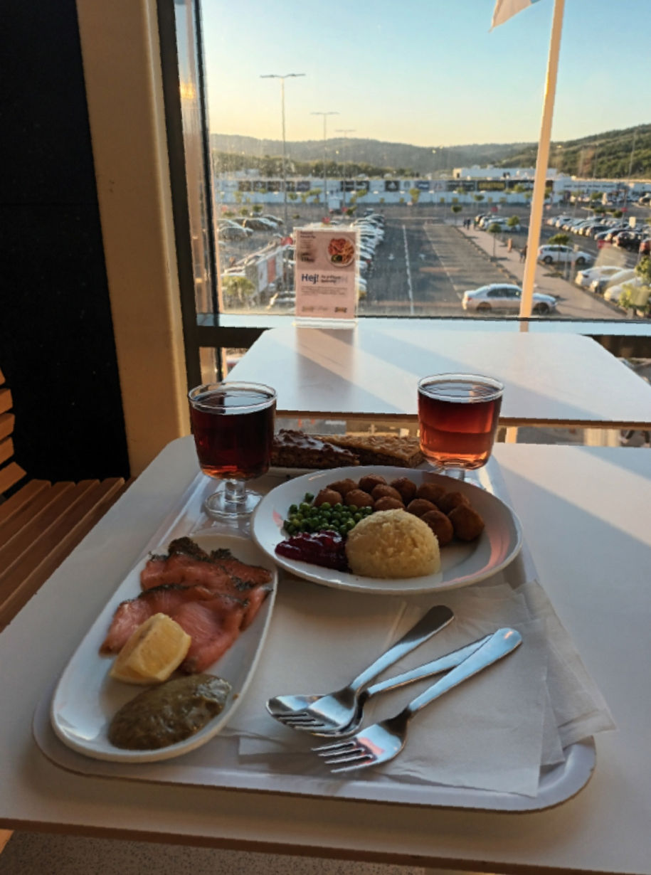 📷 Столик ресторана IKEA (логотип «Hej»): на подносе — две тарелки (лосось с лимоном; шведские тефтели с пюре, горошком и брусничным вареньем) и два стакана с напитком; за окном — парковка, торговый центр, закат над холмами.