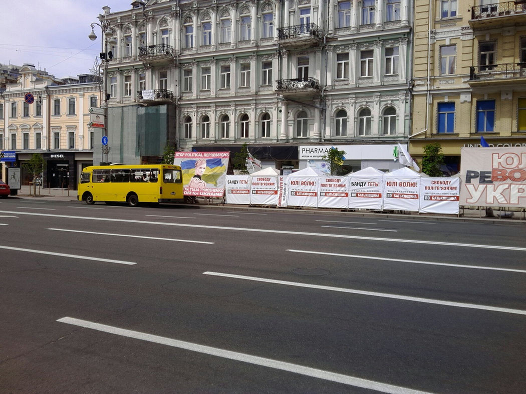 Жёлтый городской автобус у тротуара с рядом белых протестных палаток с лозунгами «Банду геть!», «Юлі волю!», «Stop political repressions»