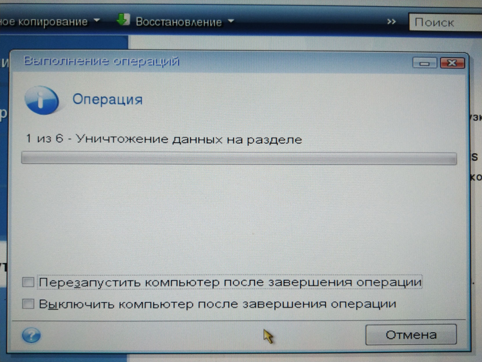 Скриншот программы Acronis True Image (Next Generation): диалоговое окно «Выполнение операции», заголовок «Операция»; строка прогресса «1 из 6 - Уничтожение данных на разделе» (~50% заполнения); чекбоксы «Перезапустить компьютер после завершения операции» и «Выключить компьютер после завершения операции»; кнопка «Отмена»; синий фон окна — фото с монитора под углом
