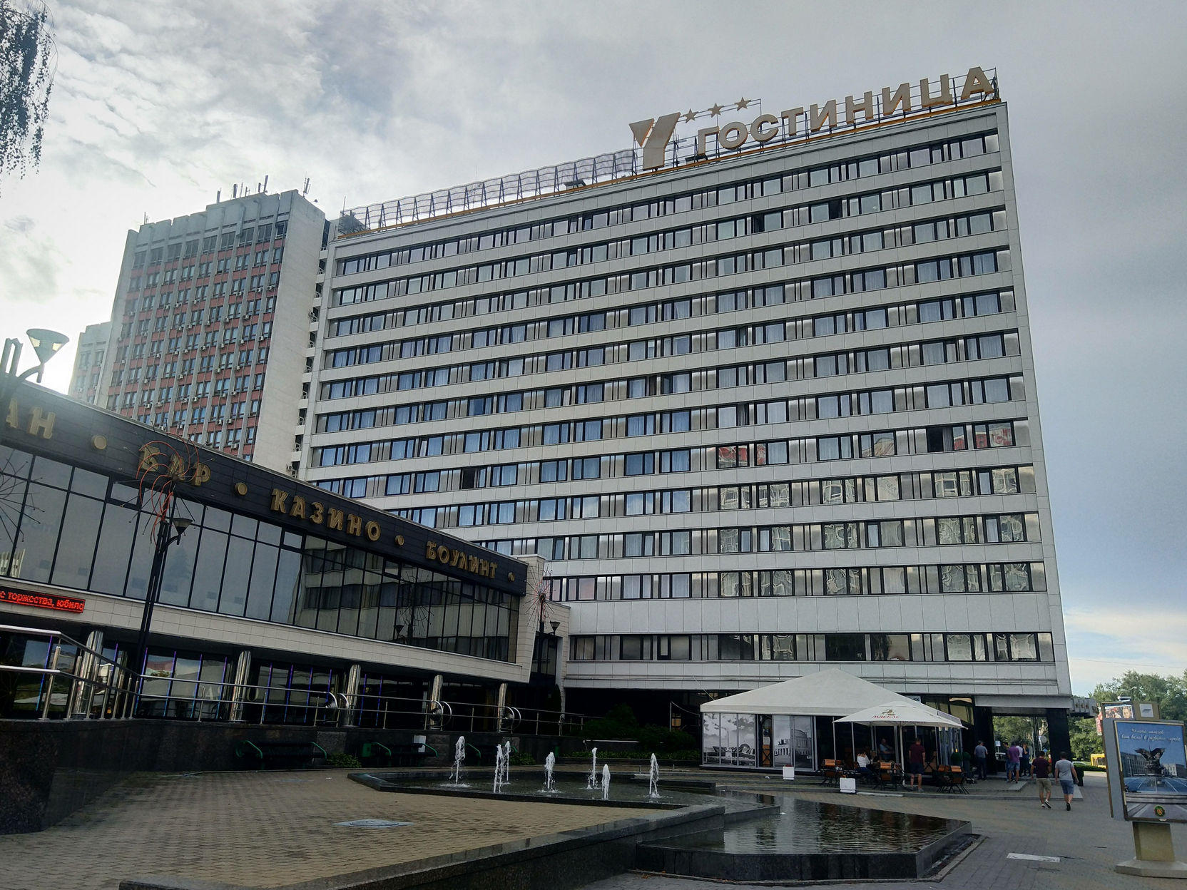 Фасад советской гостиницы «Юбилейная» в Минске: многоэтажное здание позднего советского модернизма — светло-серый бетонный фасад с рядами прямоугольных окон; на крыше надпись «ГОСТИНИЦА»; на нижнем уровне пристройки вывеска «БАР · КАЗИНО»; перед входом небольшая площадь с фонтаном; пасмурное небо, зелёные деревья