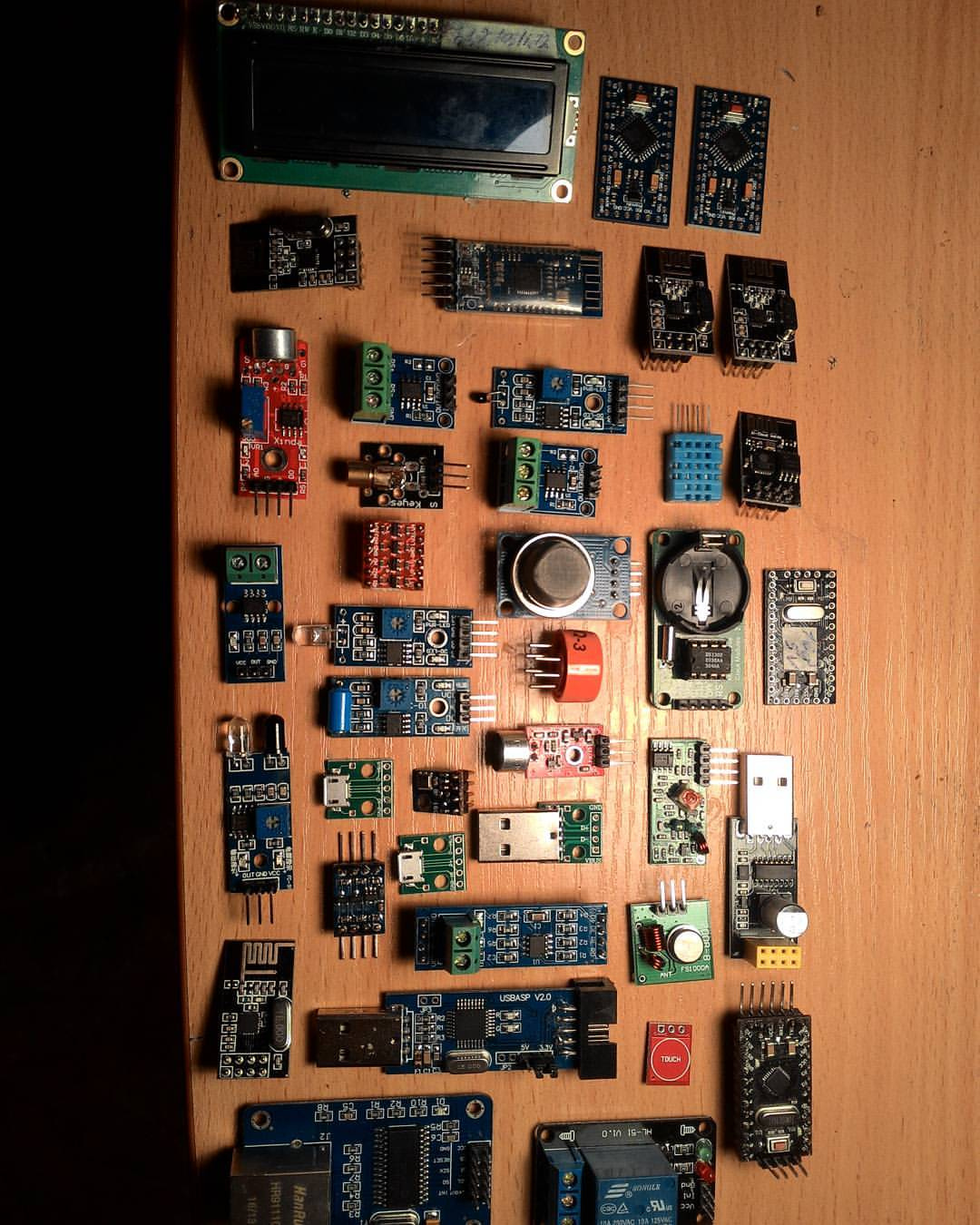 Вид сверху: около 40 Arduino-совместимых электронных модулей разложены рядами на деревянной столешнице — LCD-дисплей (16×2), датчики температуры и влажности (DHT), датчики газа, ИК-приёмники, реле, Bluetooth-модули, Arduino-платы (Nano, Mini), модули беспроводной связи, часовые модули RTC и другие небольшие PCB-платы