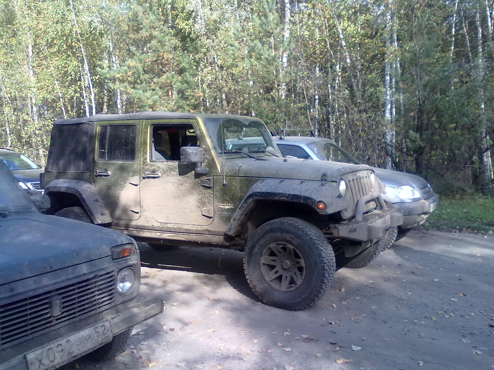 Грязный Jeep Wrangler на грунтовой стоянке среди берёз и сосен