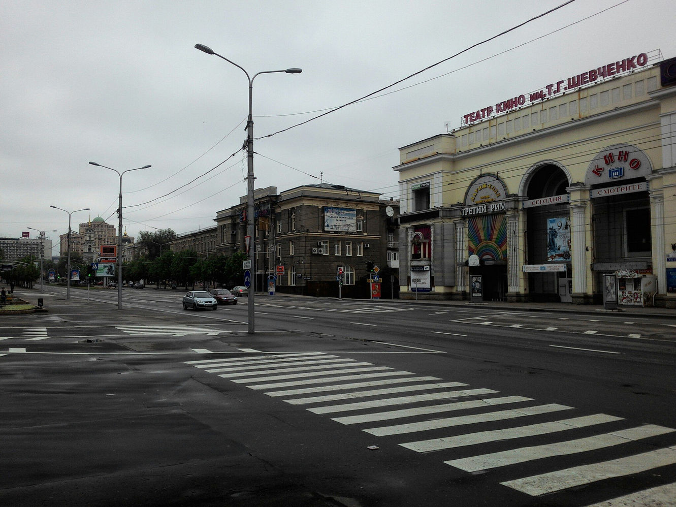 Пасмурный день в Донецке: перекрёсток с пешеходным переходом; справа — здание театра кино им. Т.Г. Шевченко (афиша «Третий Рим»); вдали купола церкви; машины на мокром асфальте