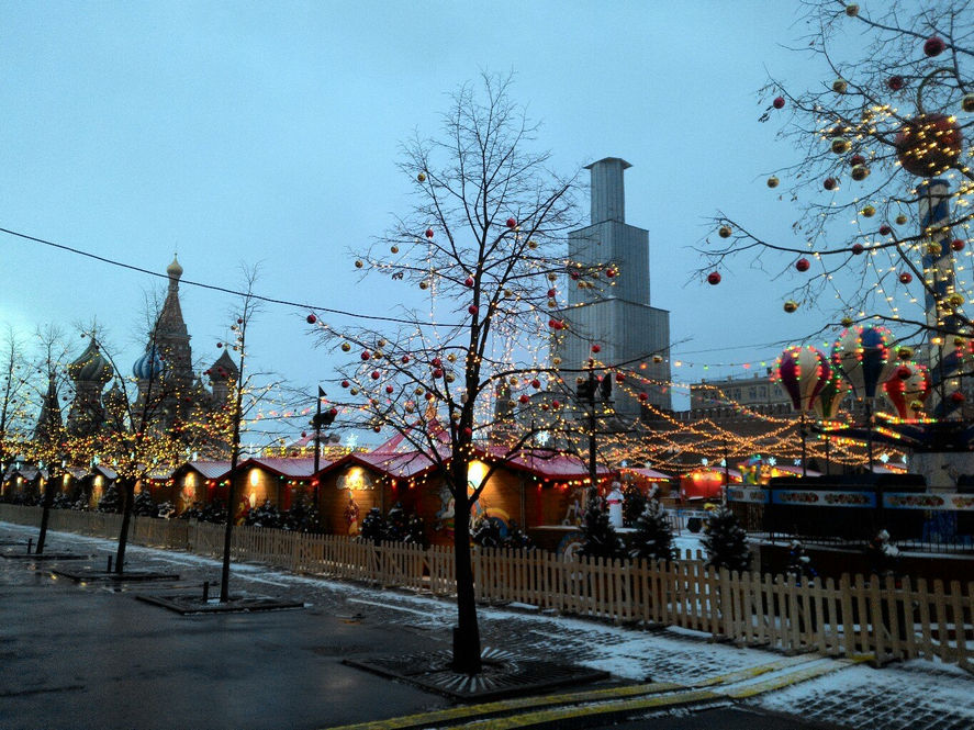 Красная площадь в Москве: новогодняя ярмарка — деревянные домики с красными крышами, деревья украшены цветными шарами и гирляндами, разноцветные воздушные шары-аттракционы; вдали — купола храма Василия Блаженного; пасмурный серый зимний день