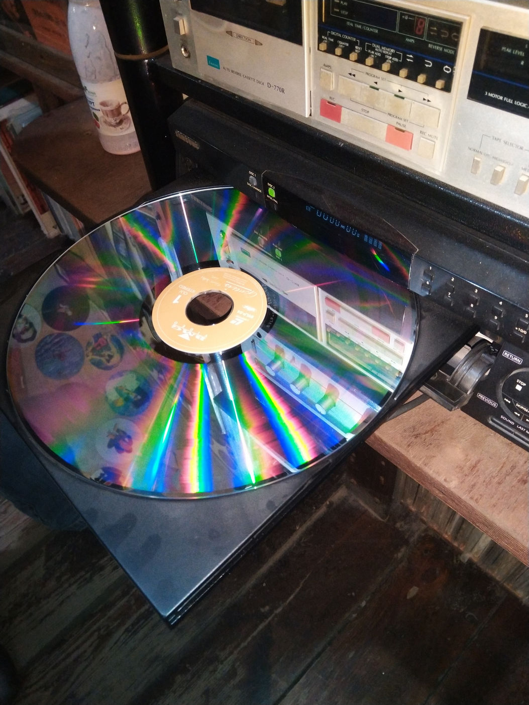 Крупным планом большой лазерный диск (LaserDisc) на лотке проигрывателя; поверхность переливается радужными цветами (зелёный, синий, фиолетовый, золотистый); на заднем плане — ретро-аппаратура с оранжевыми индикаторами и кнопками управления