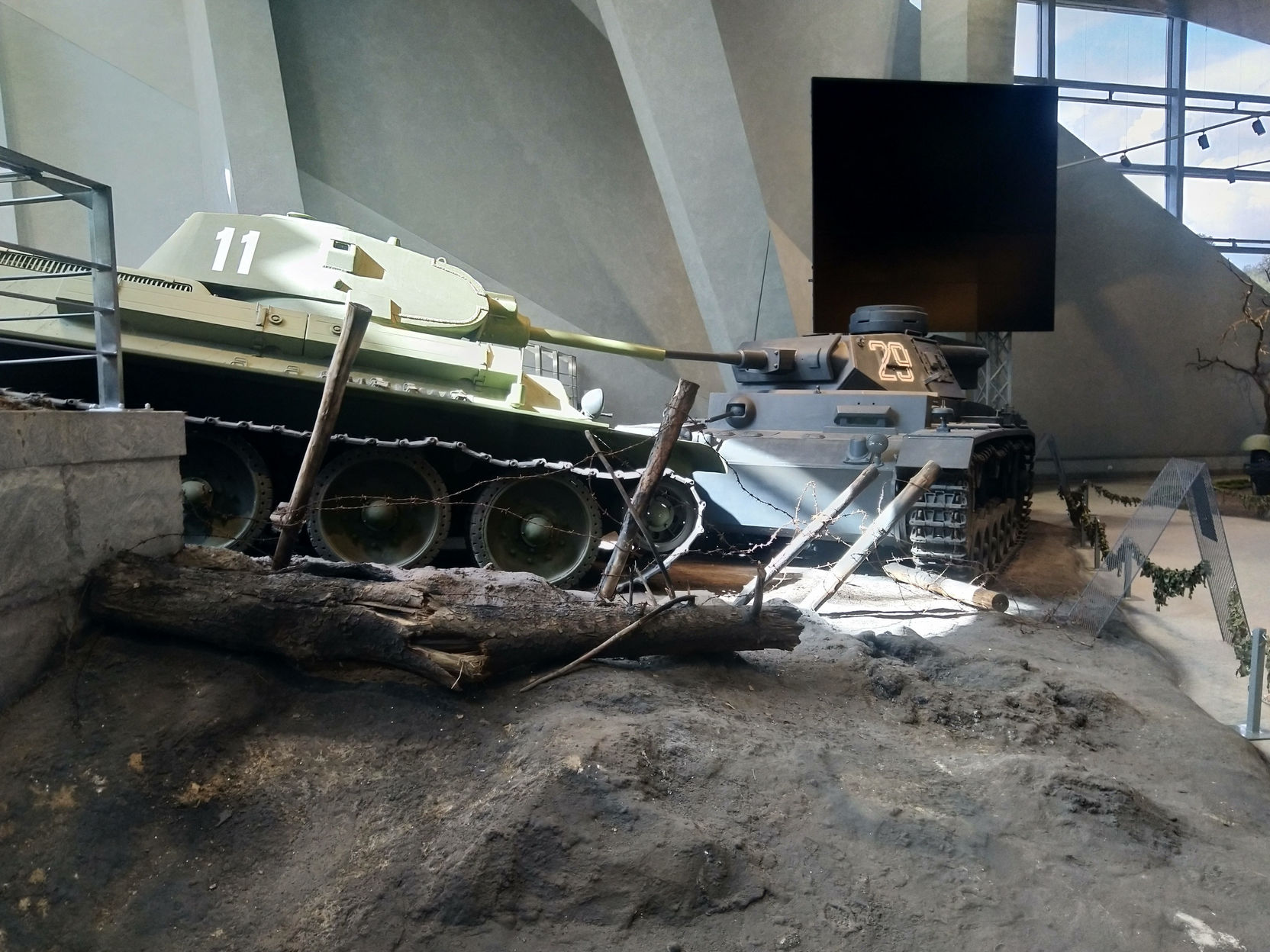 Танк Т-34 с бортовым номером «11» в зелёном камуфляже в музейной диораме: под гусеницами грунт и брёвна противотанковых заграждений, натянута цепь; справа фрагмент светлой техники (возможно немецкой); светлые стены современного музейного здания с большими окнами и лестницей на заднем плане