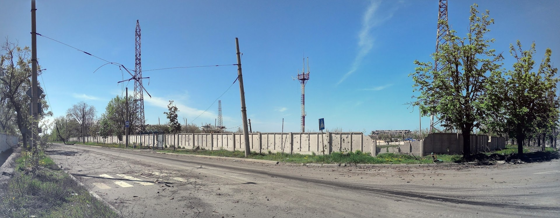 📷 Панорама разрушенного украинского города: широкая пустая улица, бетонный забор, покосившиеся электроопоры, деревья, голубое небо.