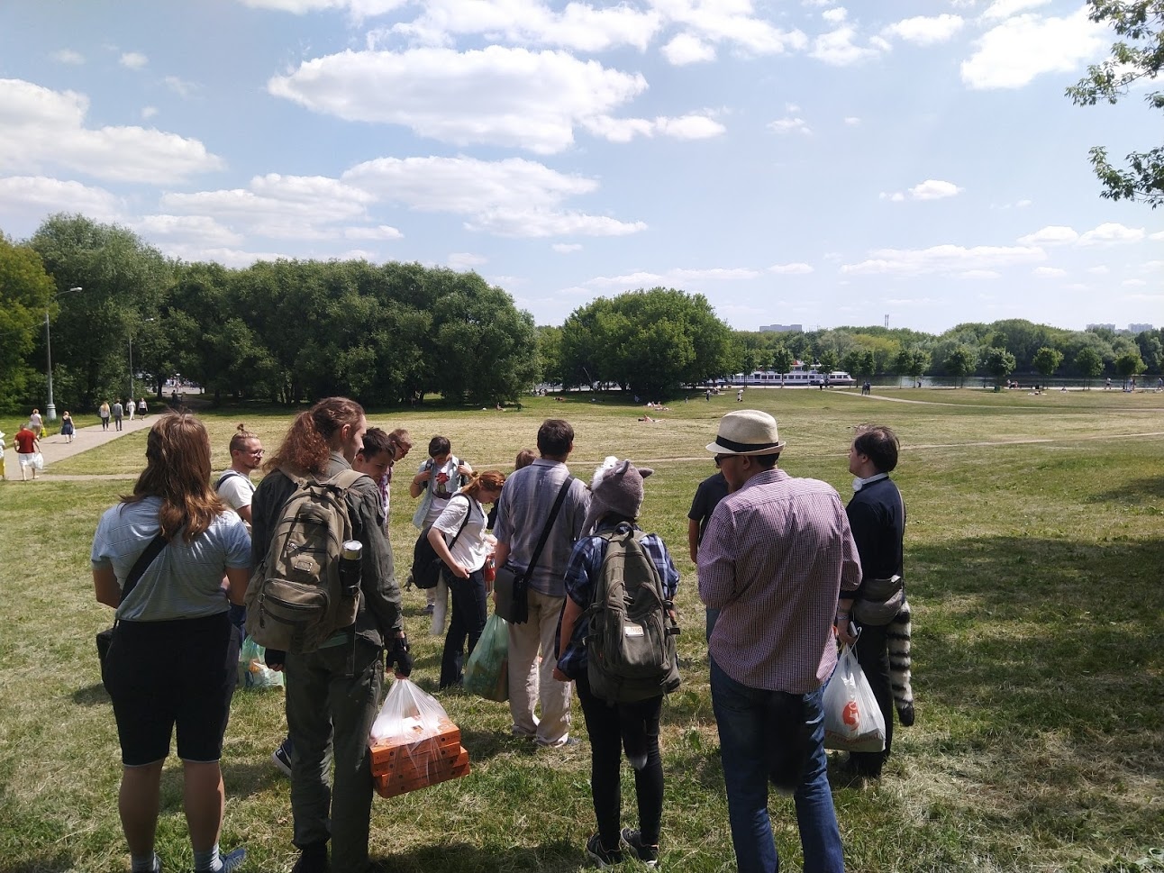 📷 Группа людей с рюкзаками стоят в солнечном парке, смотрят на открытое поле с деревьями вдали.
