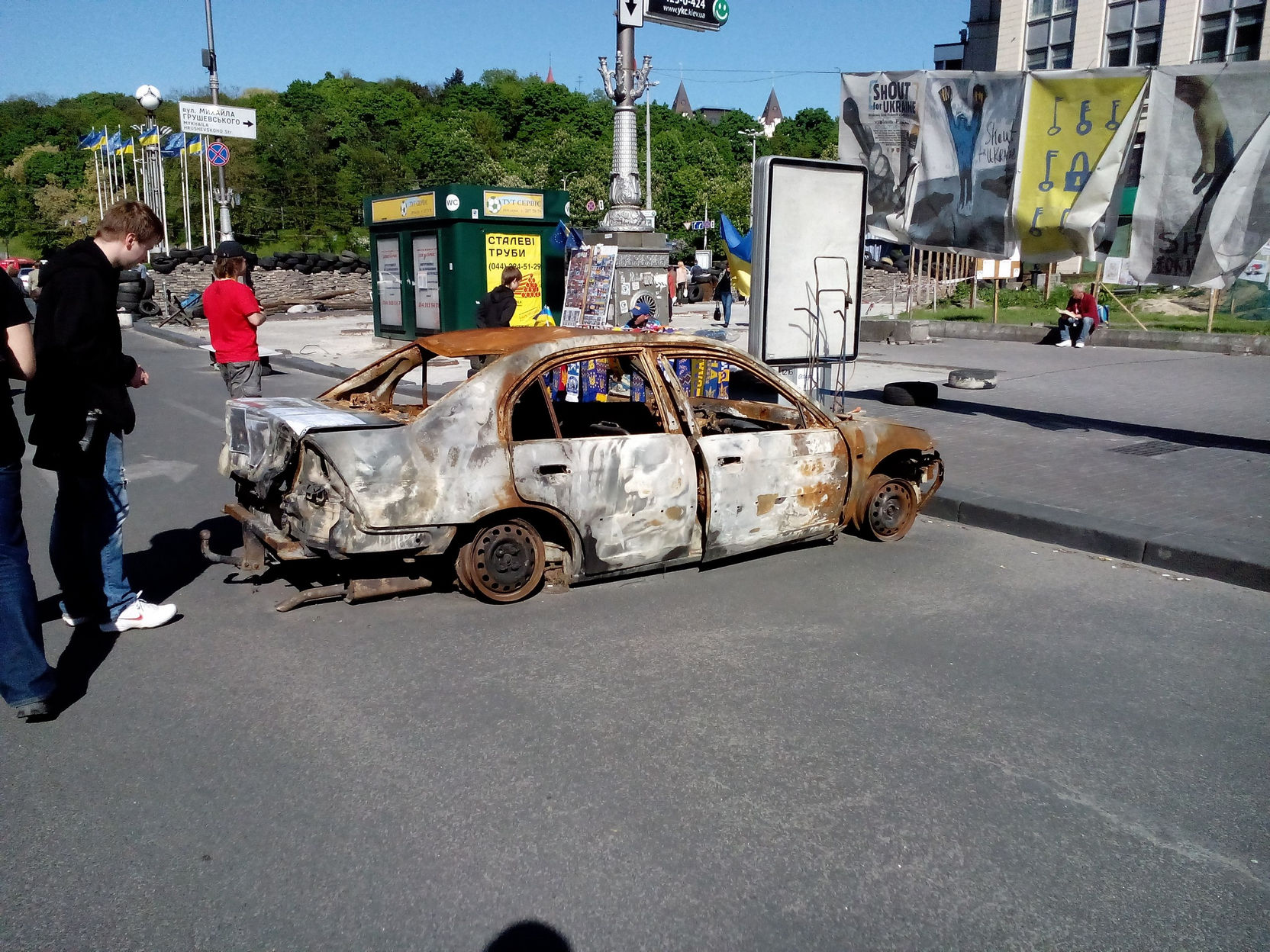 Сгоревший до каркаса автомобиль посреди улицы у Майдана; прохожие идут рядом, на заднем плане — украинские флаги и баннеры