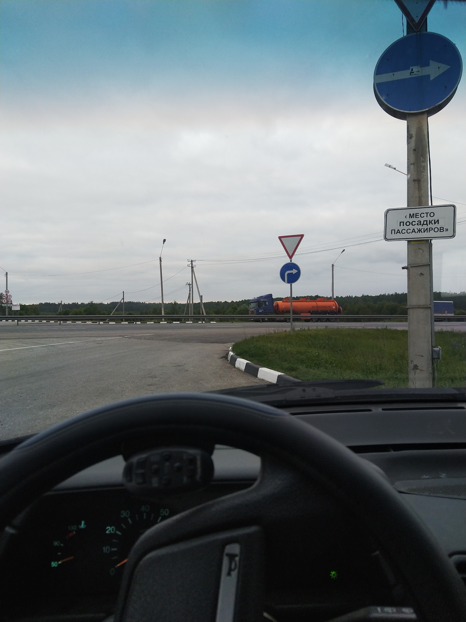 Вид из салона автомобиля через лобовое стекло на перекрёсток загородной дороги: столб с дорожными знаками — синий круглый «обязательное направление направо», треугольный «уступи дорогу», синий круглый «круговое движение», прямоугольная табличка «МЕСТО ПОСАДКИ ПАССАЖИРОВ»; вдали на трассе — оранжевая цистерна-грузовик; лесополоса по горизонту; пасмурное серо-белое небо; руль и тёмная приборная панель на переднем плане
