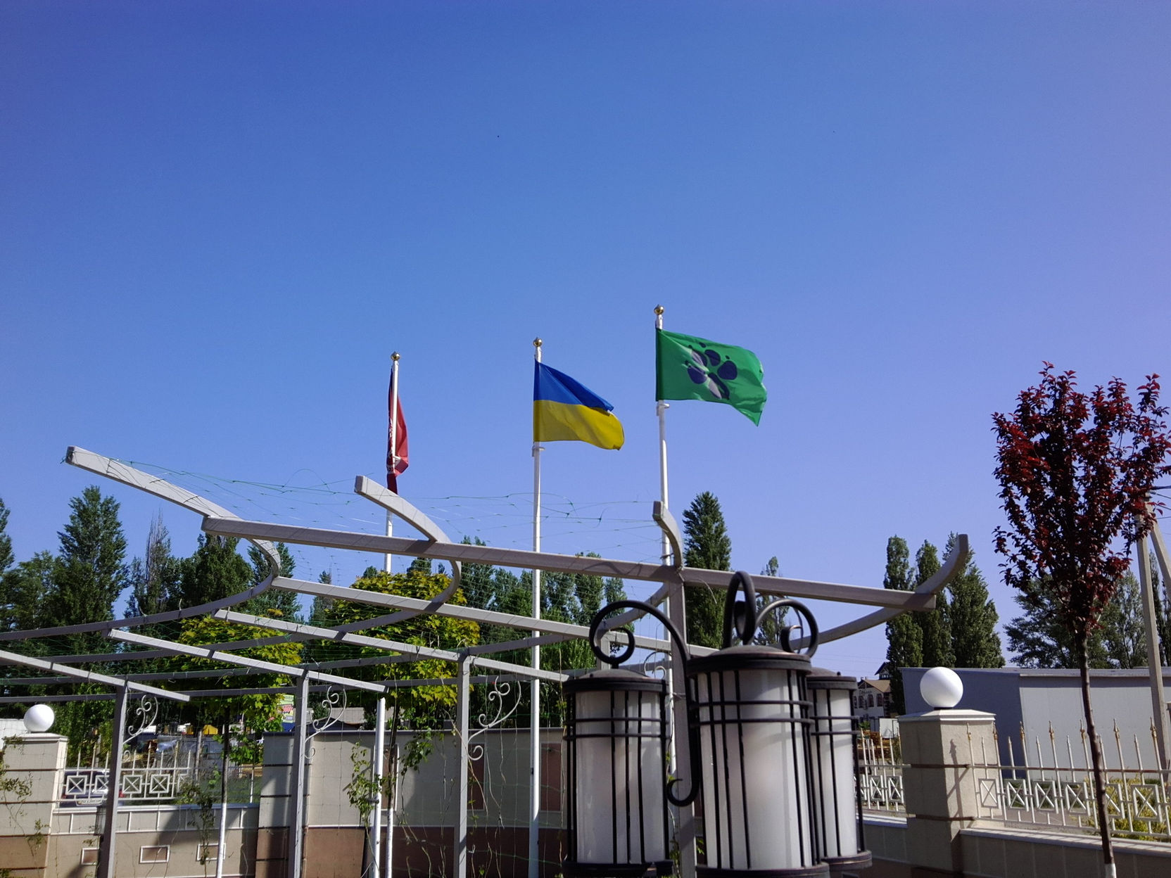 Три флага на флагштоках у входной арки: красный, украинский сине-жёлтый и зелёный с символом — на фоне ясного синего неба