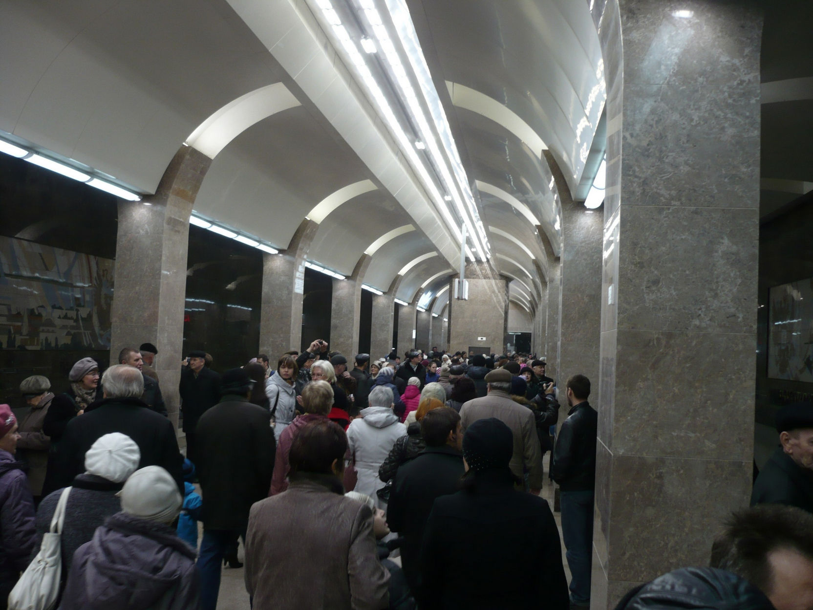 Платформа новой станции метро в день открытия: толпа горожан в зимней одежде идёт по перрону; арочный потолок с люминесцентными светильниками, колонны и стены отделаны серым мрамором